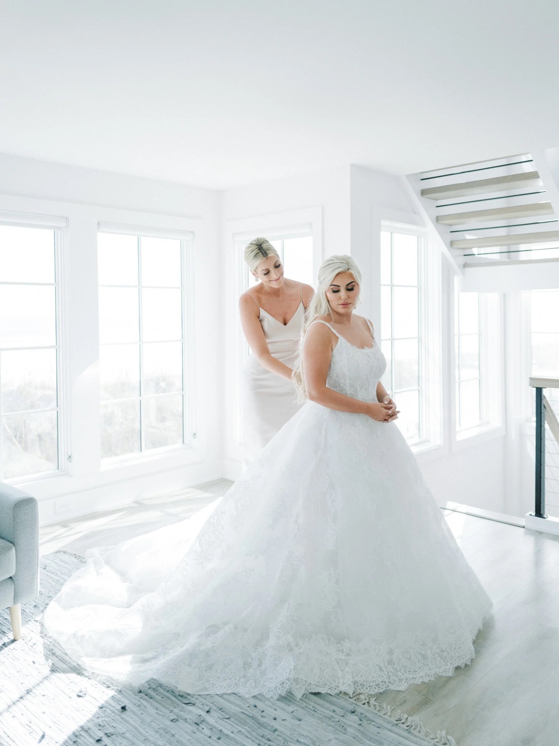 A bride in a wedding gown standing in a bright white room with large windows, while another woman helps her with her dress.