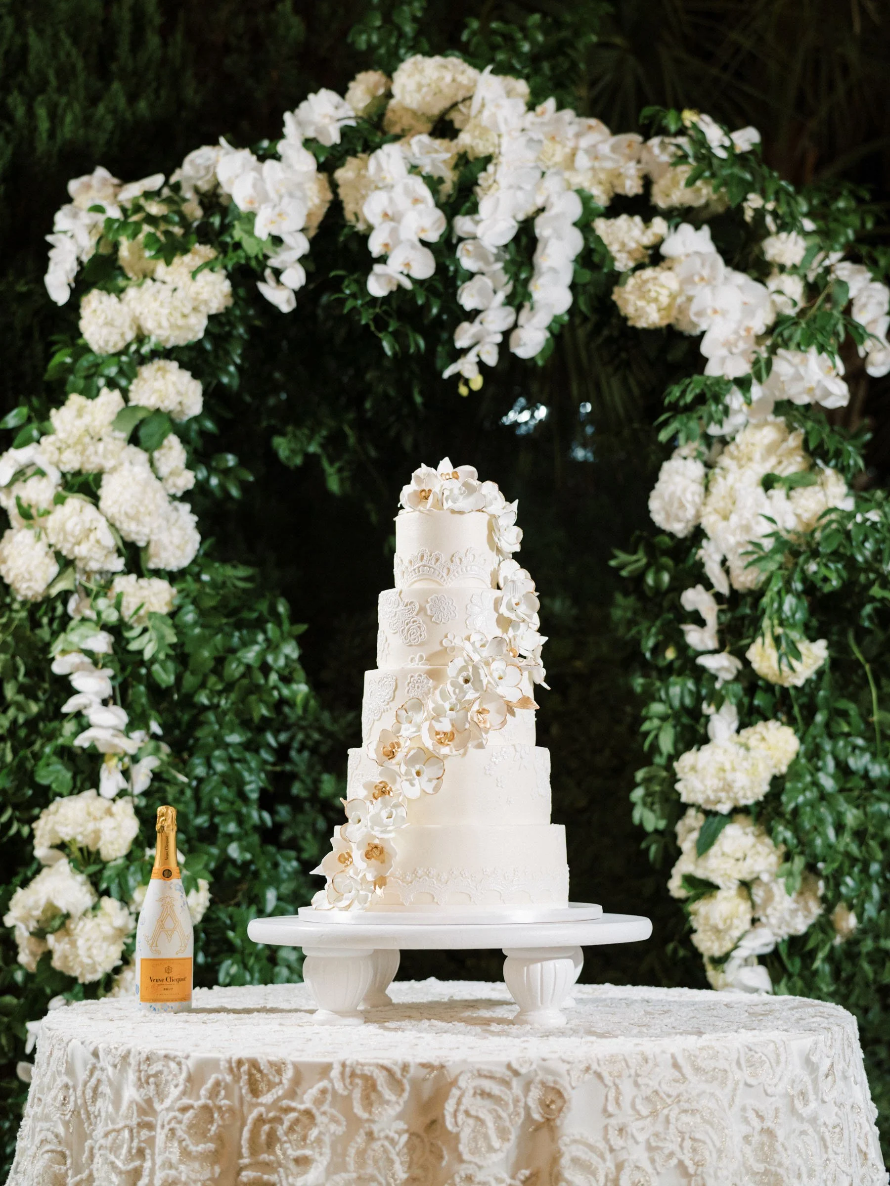 A tall, white wedding cake decorated with white flowers on a cake stand, on a round table covered with a lace tablecloth, with a floral arch made of white flowers and greenery in the background, and a bottle of Veuve Clicquot champagne on the table.