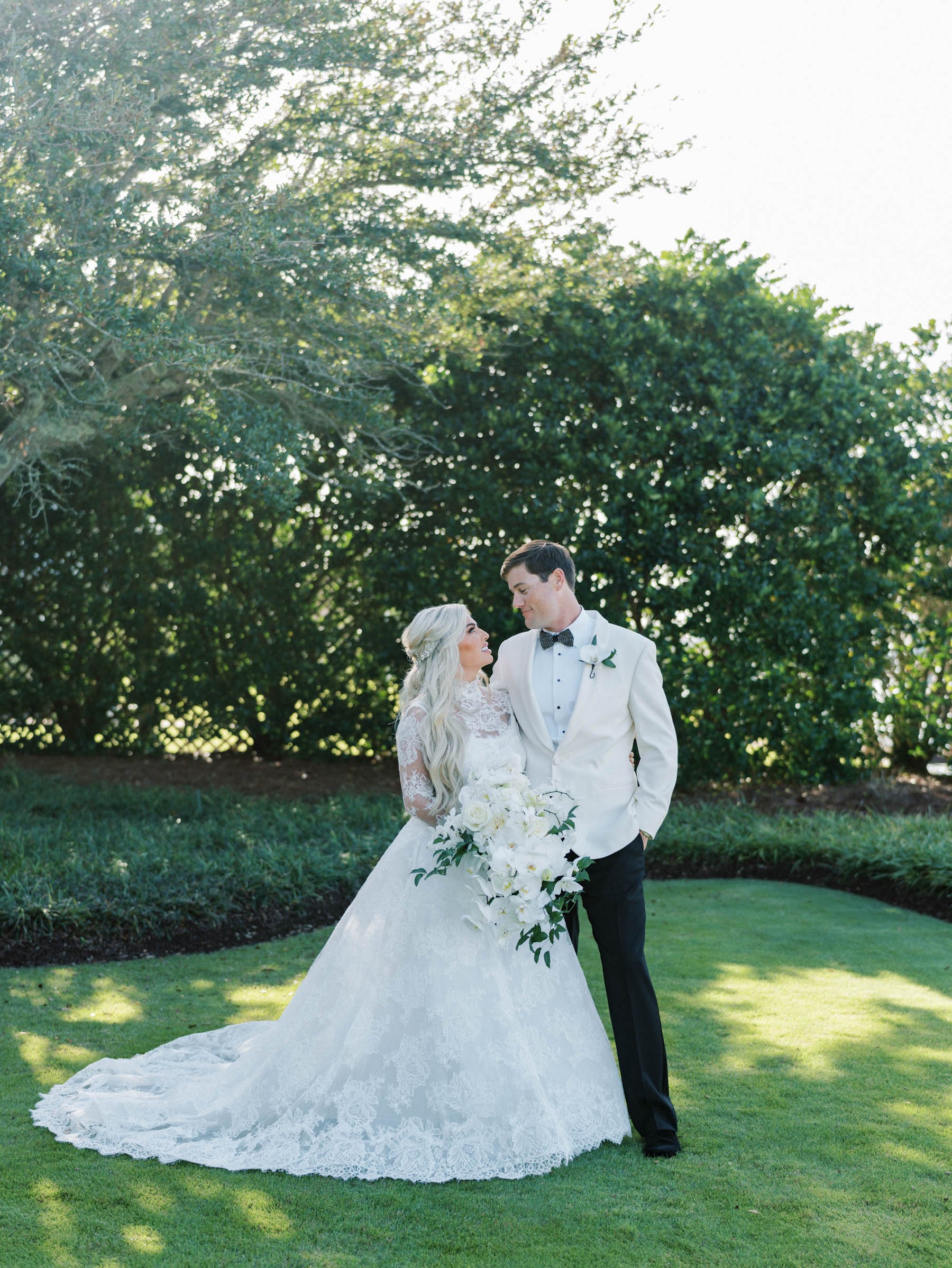 A bride and groom standing together outdoors, gazing at each other, with the bride holding a large bouquet of white flowers, on a green lawn with trees in the background.