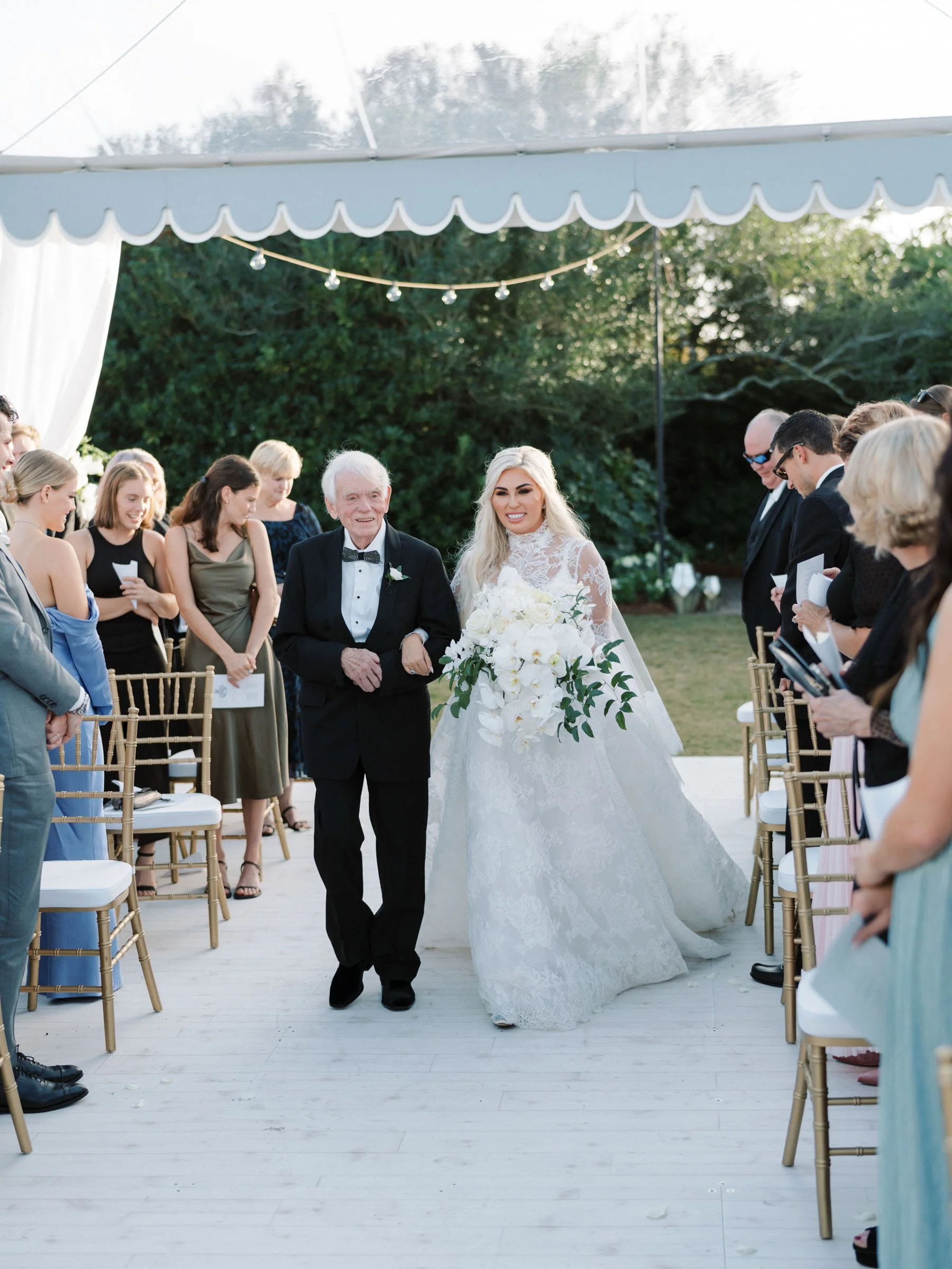 A bride walking down the aisle at her outdoor wedding, accompanied by an older man, likely her father, surrounded by seated and standing guests.