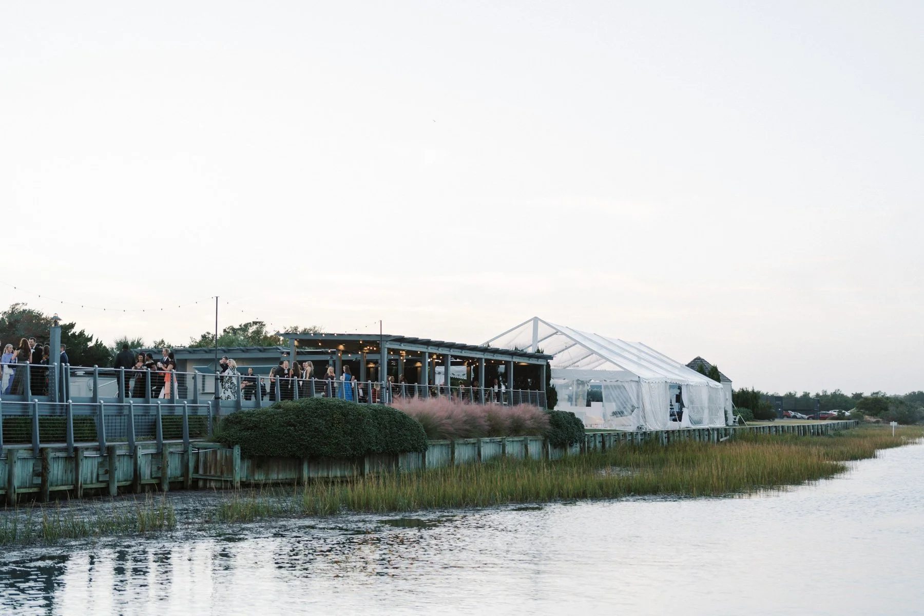 People gathered on a waterfront deck during an outdoor event at sunset, with a large tent nearby.