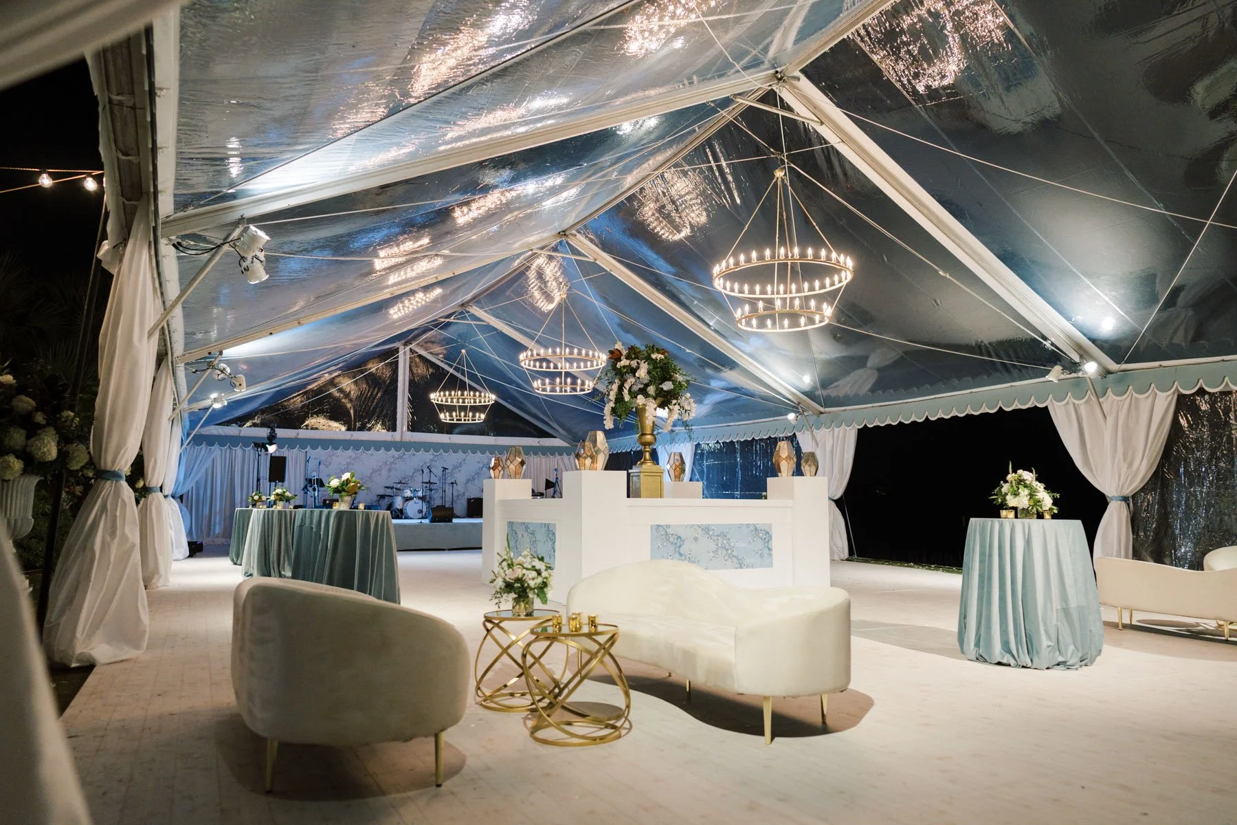 Elegant event tent with chandeliers, floral arrangements, and decorated tables, set up for a celebration or wedding reception at night.