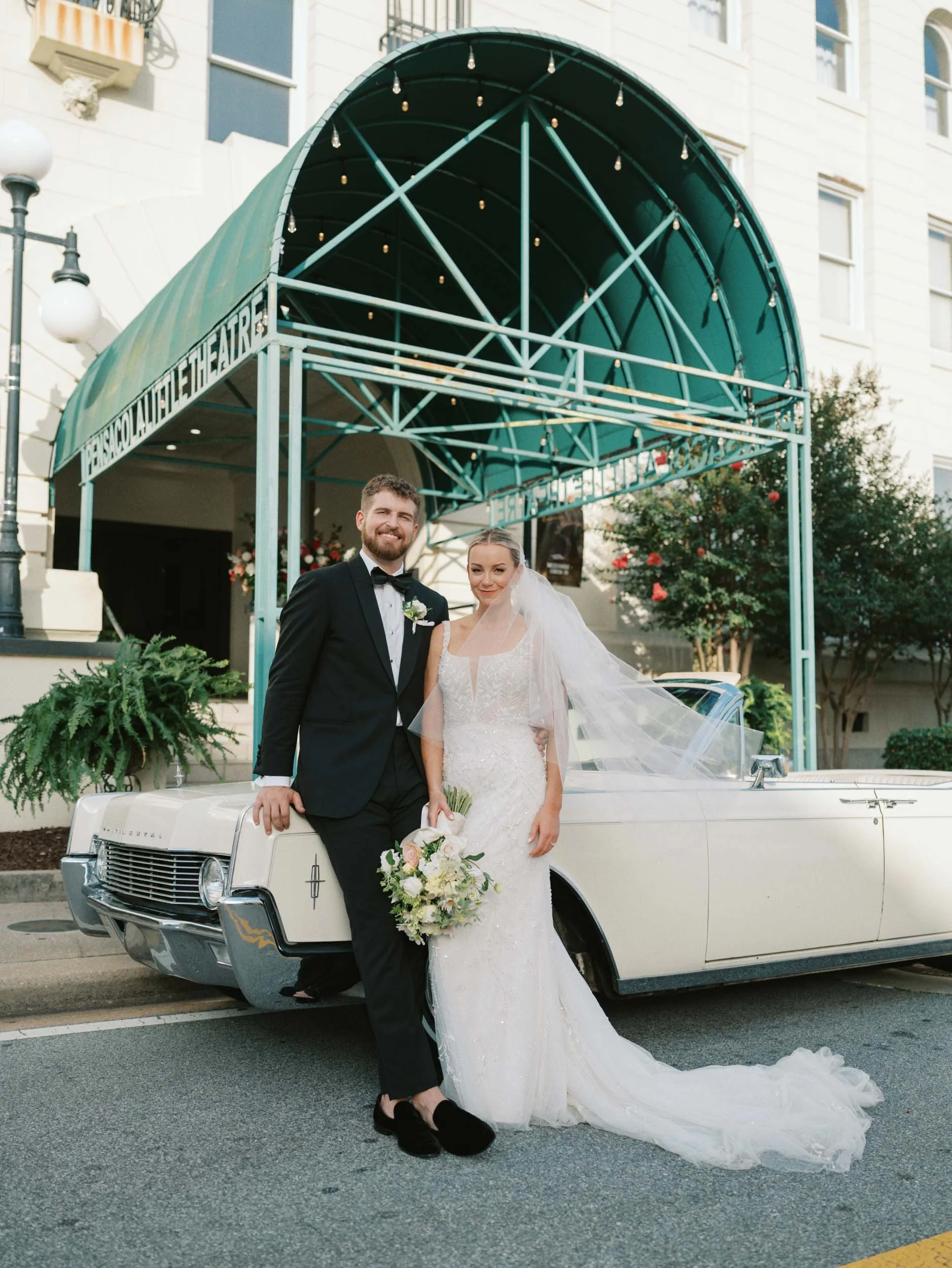 A bride and groom in wedding attire standing in front of a vintage white car, posing for a photo outside a theater with a sign that reads "Pascal Theatre".