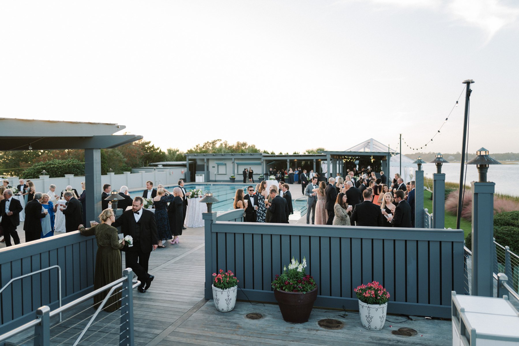 Guests at an outdoor wedding reception on a deck overlooking a lake, dressed in formal attire, with a pool and a tent in the background, and string lights hanging above.