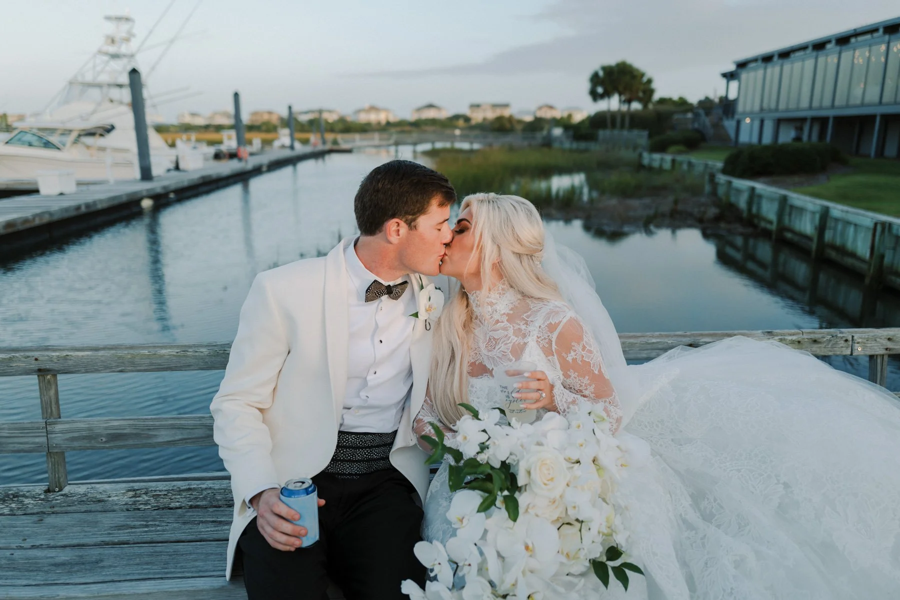 Bride and groom sharing a kiss on a dock near water, with boats and a building in the background, during sunset.