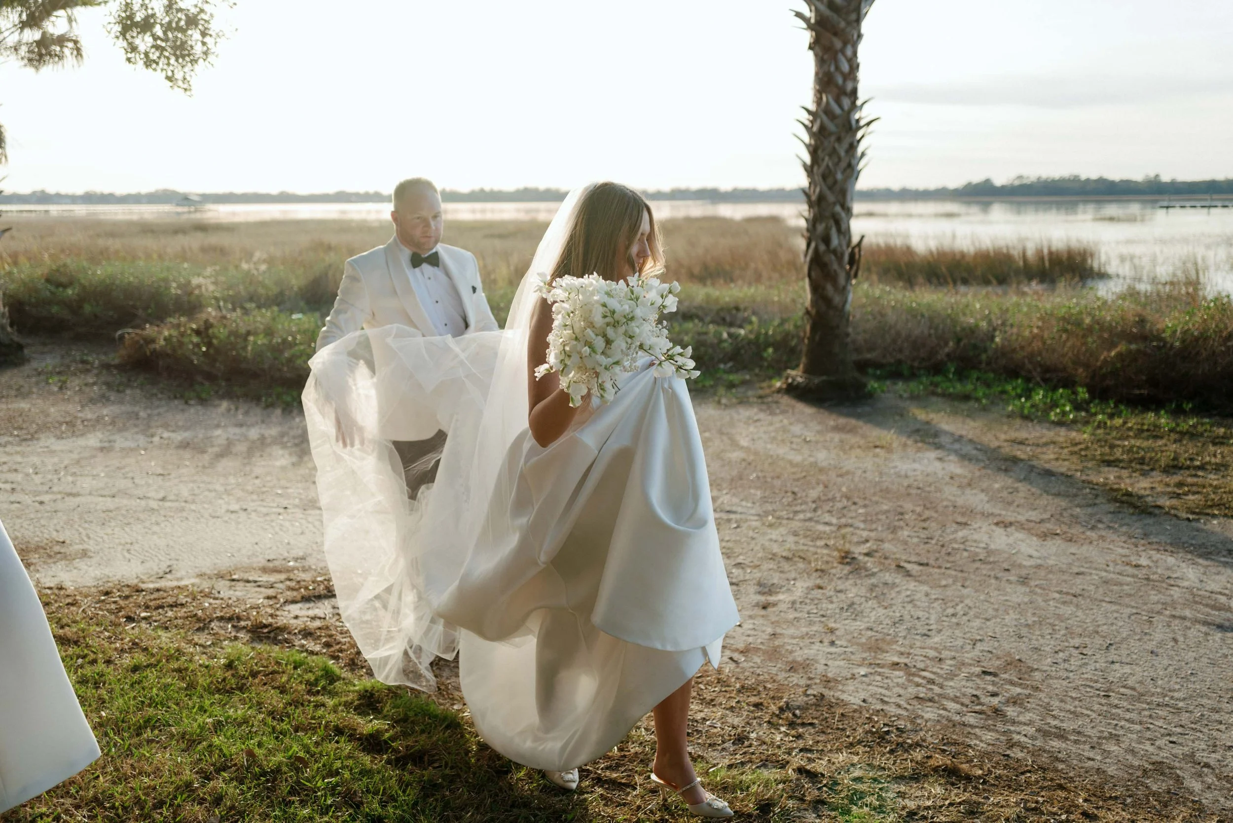 Bride holding a bouquet arriving at outdoor wedding on a dirt path by water with groom following behind, trees on each side, during golden hour.