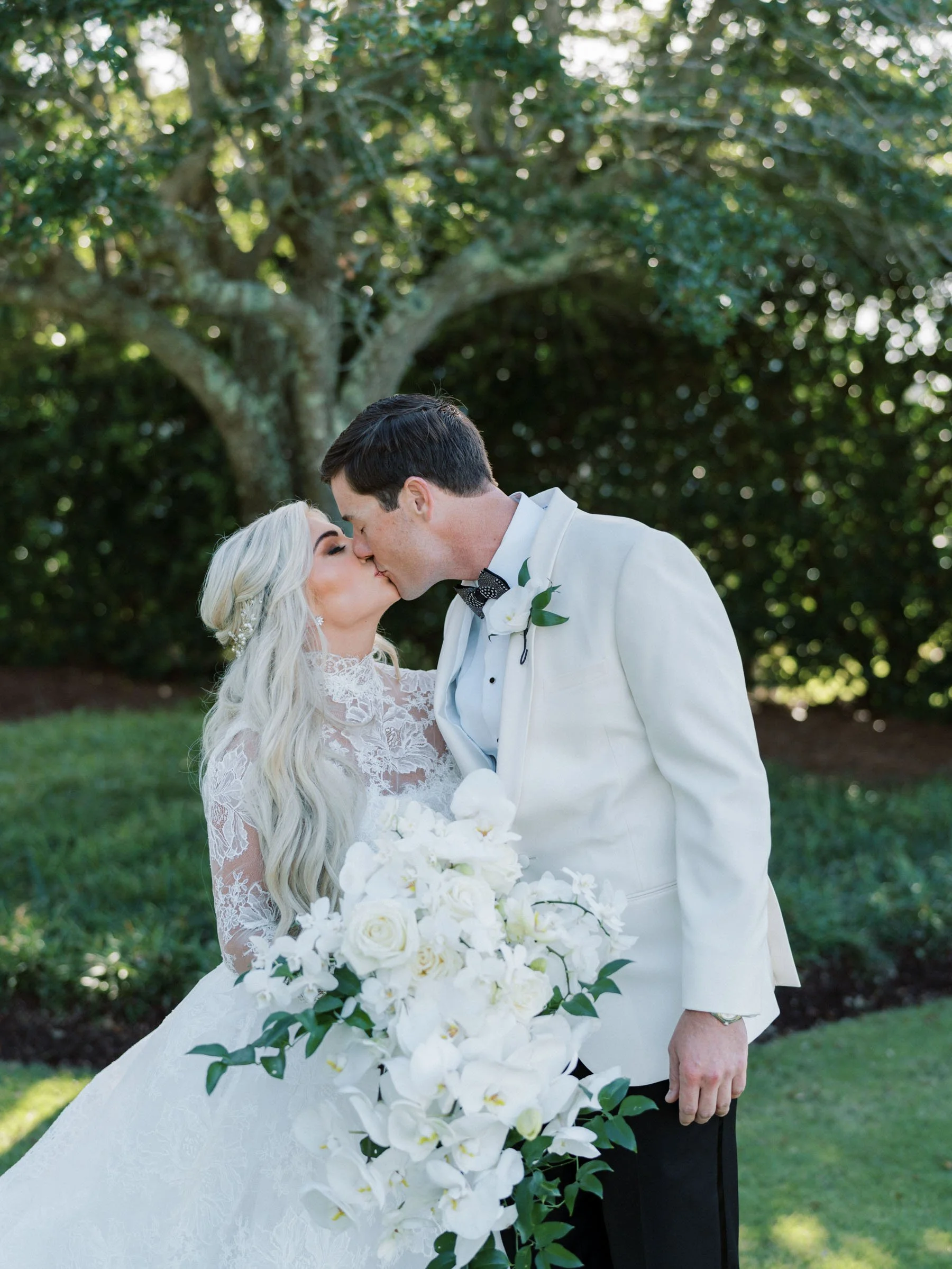 A bride and groom sharing a kiss outdoors during their wedding, with the bride holding a large bouquet of white flowers and greenery, and a large tree in the background.
