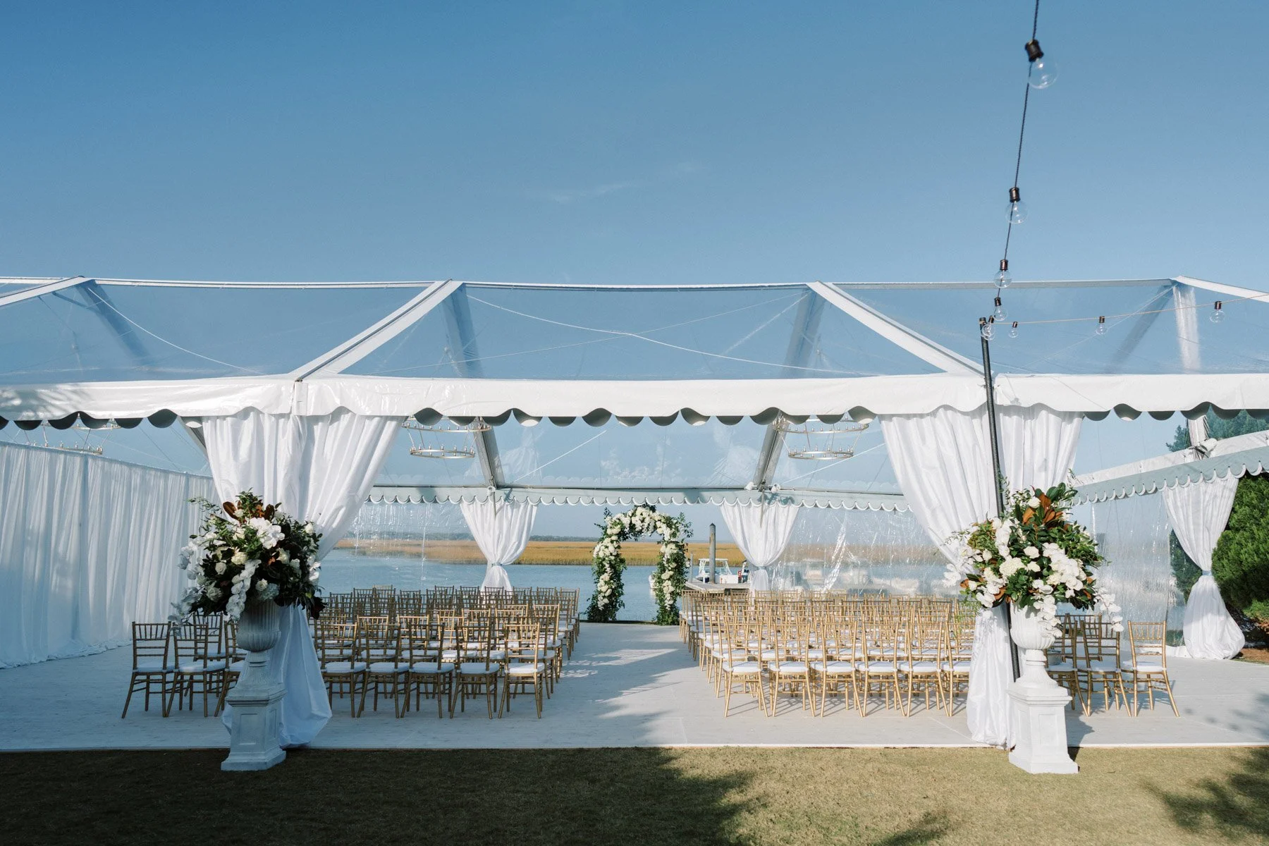 Outdoor wedding ceremony setup under a white tent with a view of water and mountains, decorated with floral arrangements and an arch.