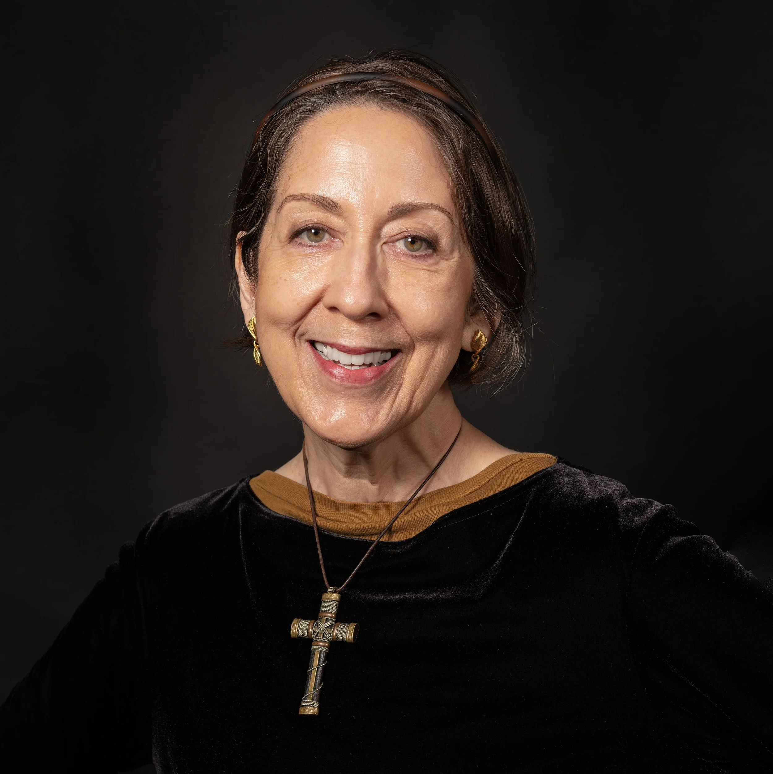 Close-up of a smiling mature woman with grayish-brown hair wearing earrings and a necklace with a cross pendant, against a black background.
