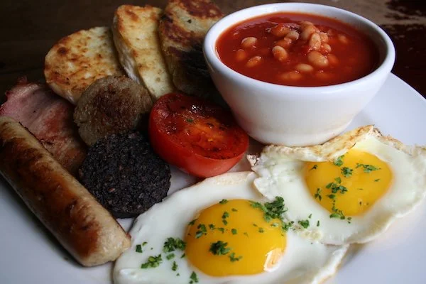Cooked breakfast with fried eggs, baked beans, sausage, bacon, black pudding, white pudding, tomato, toast, soda farl, and potato farl on a white plate.