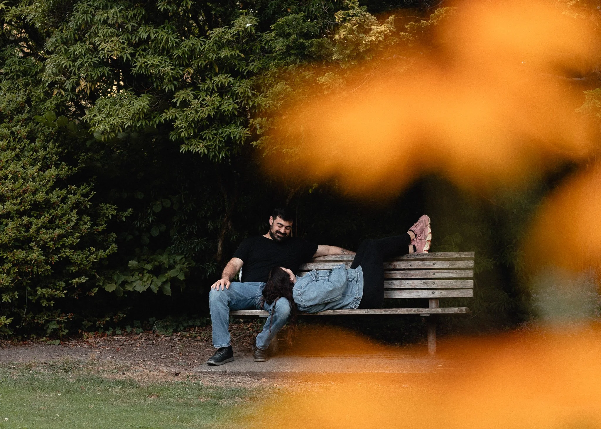 A man and a woman sitting on a park bench surrounded by green trees, with the man smiling and the woman resting her head on his lap.