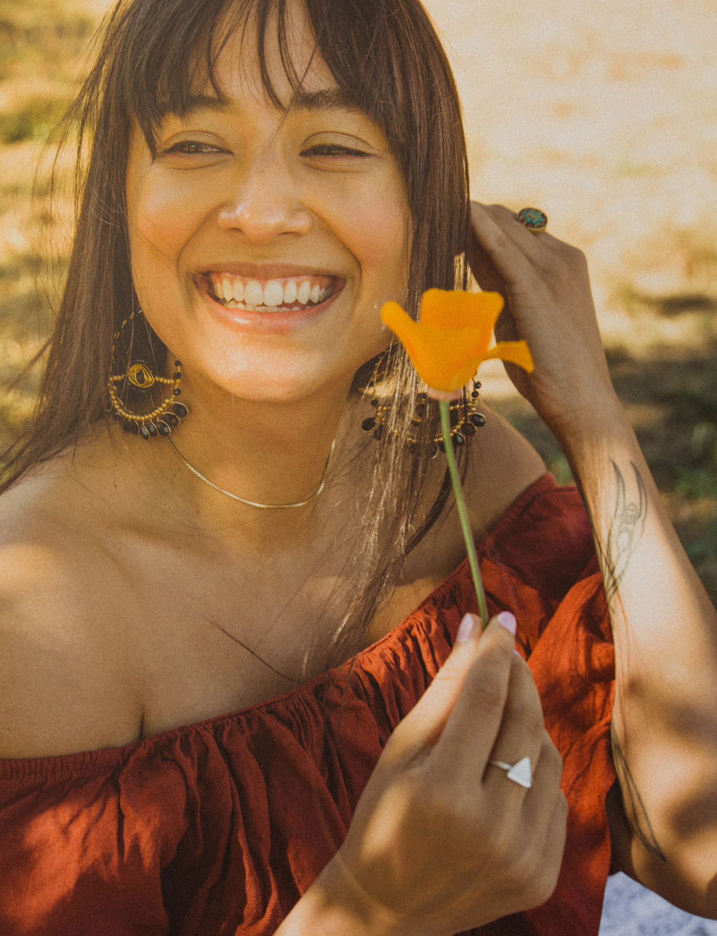 Close-up of a smiling woman holding a yellow flower, wearing an off-the-shoulder red dress and layered jewelry, with shoulder-length dark hair, outdoors in warm sunlight.
