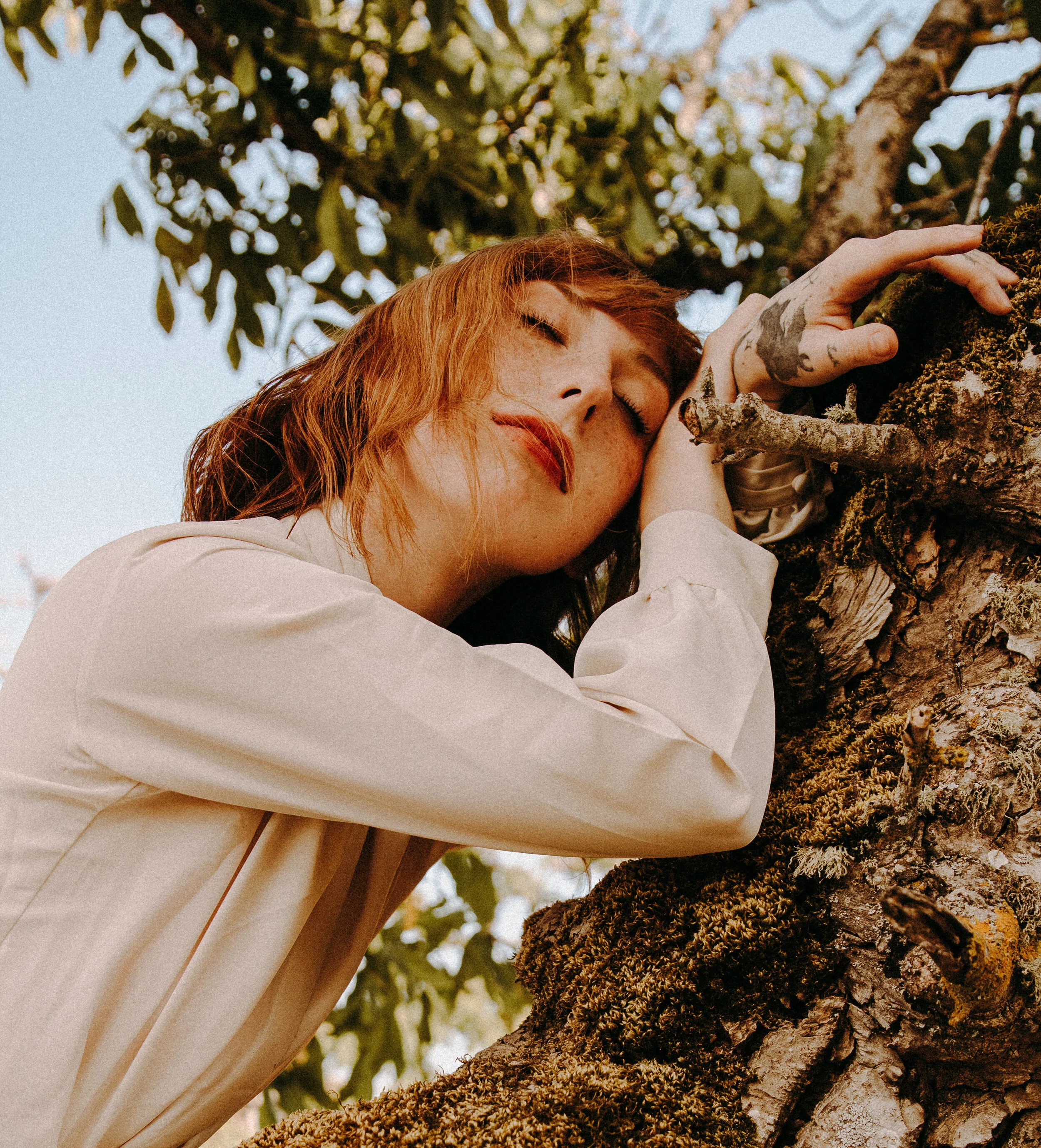 A woman with red hair and tattoos is touching moss and bark on a tree, with green leaves and sunlight in the background.