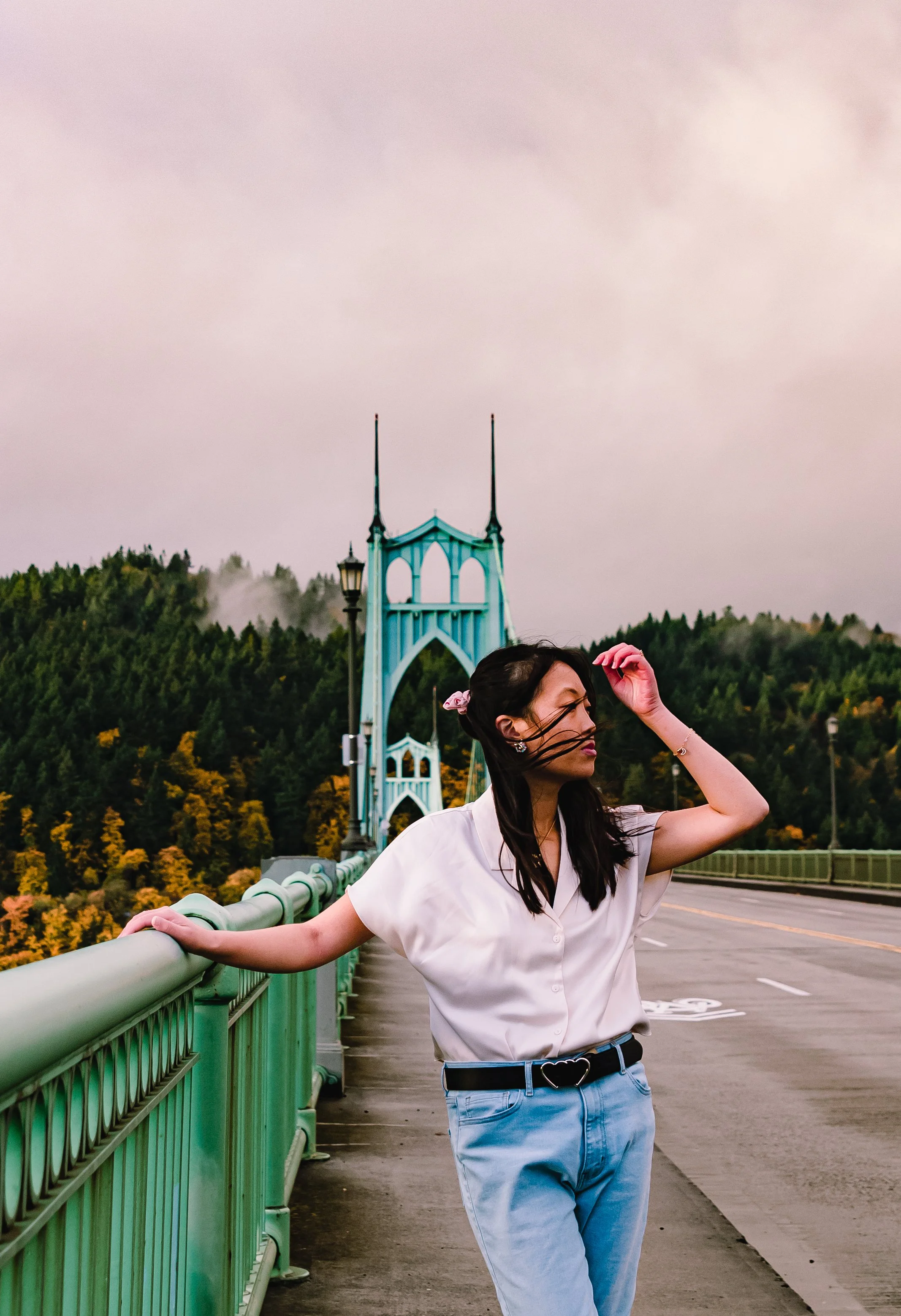A woman wearing a white shirt and blue jeans standing on a bridge with a green railing, holding onto the railing with one hand, and the bridge's scenic background features a bridge with twin spires and a lush hillside.