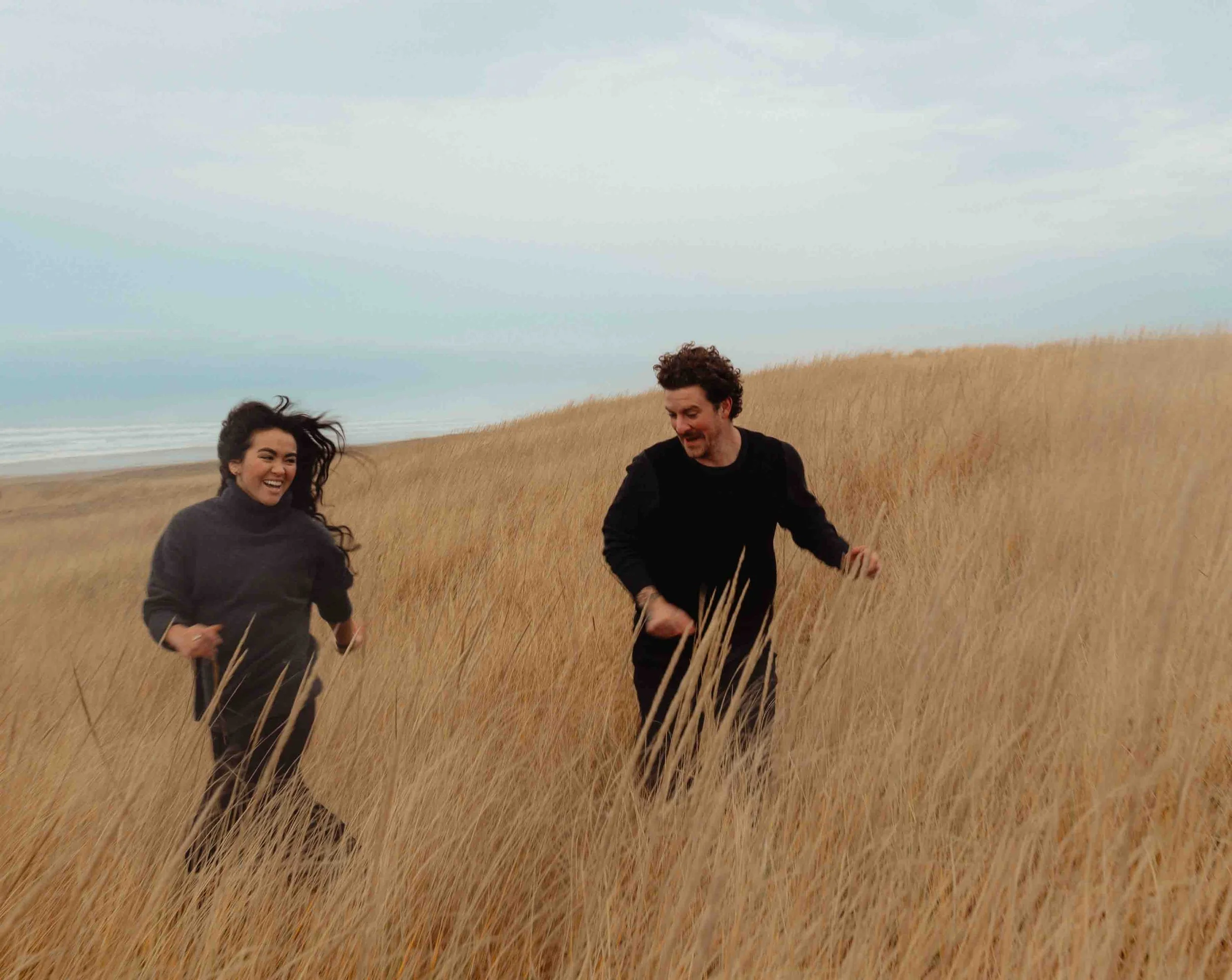 A group of people running through a field of tall, golden grass on a cloudy day.