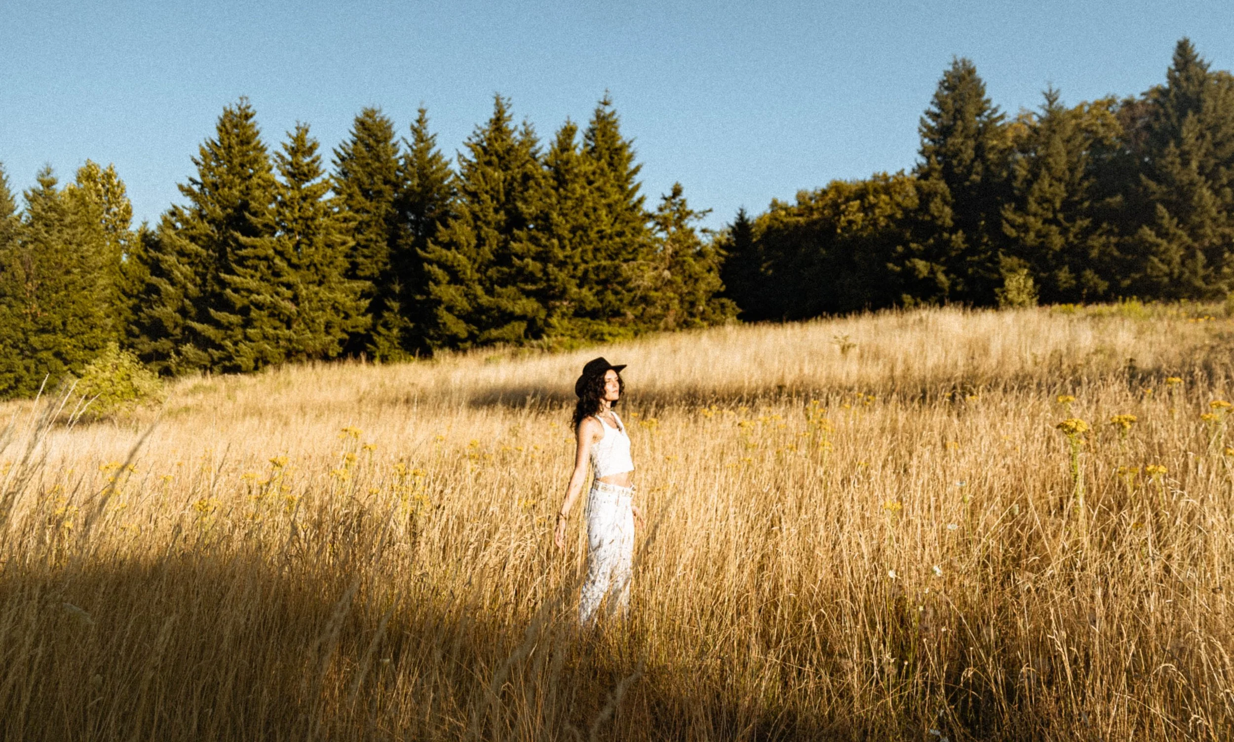 A woman with a black hat and white clothing walking through a grassy field with yellow flowers, with a background of tall evergreen trees and a clear blue sky.