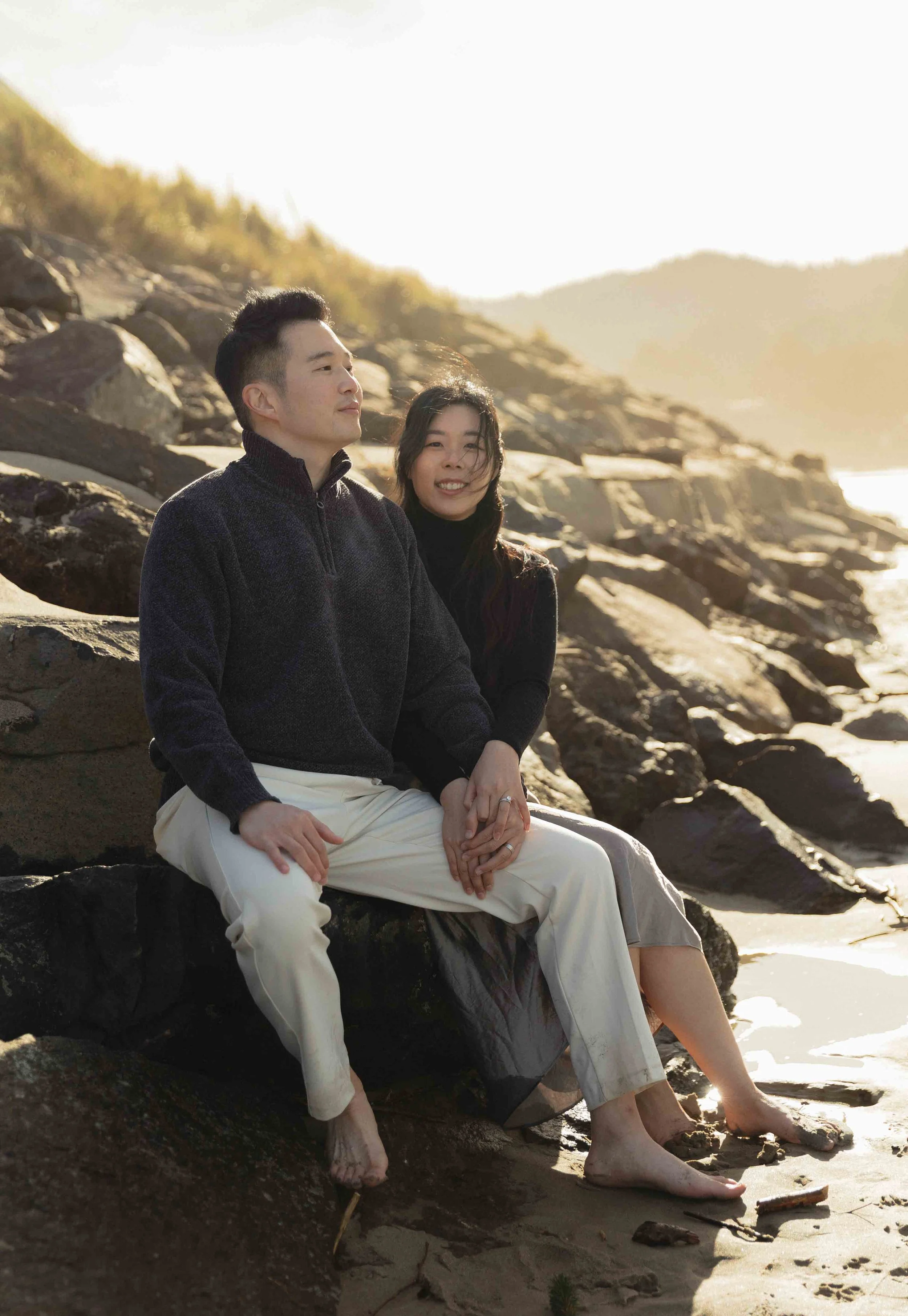 A man and woman sitting on rocks at the beach with the ocean in the background, enjoying sunset sunlight.