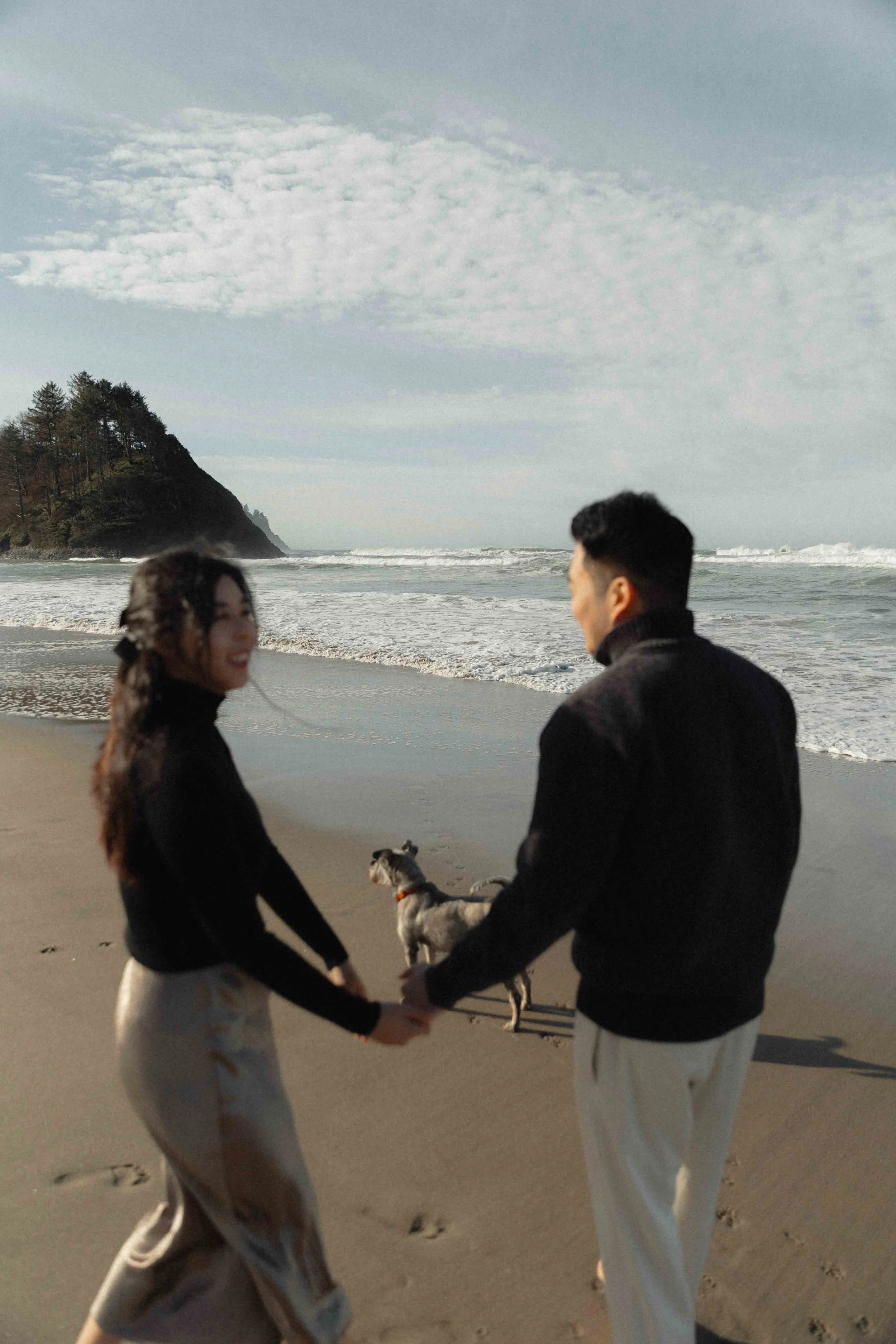 A happy couple is holding hands on a beach. The woman has long hair and is smiling, while the man has short hair. There is a dog between them, and the ocean with waves and a tree-covered hill in the background.