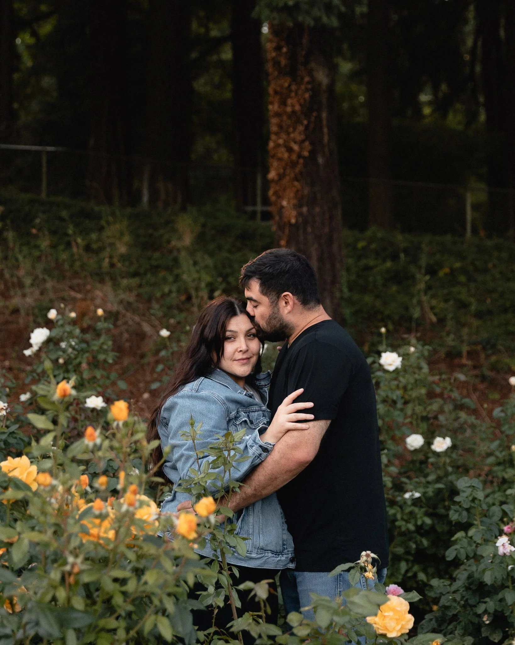 A young couple embracing in a garden filled with blooming roses. The woman has long dark hair and is wearing a denim jacket, while the man has short dark hair and is dressed in a black shirt. They are standing close together with the man's forehead g