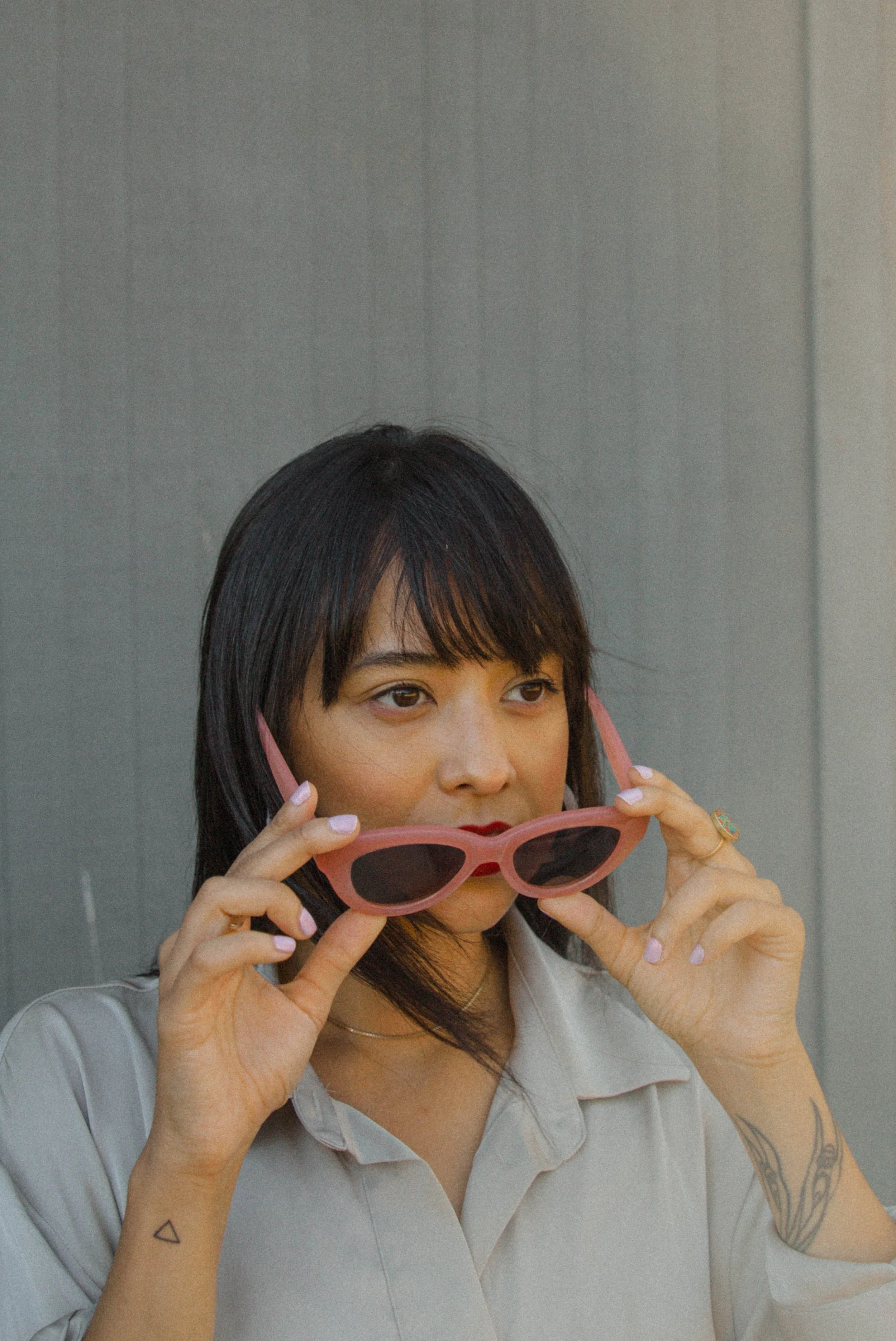Woman with dark hair and tattoos on her arms, holding pink sunglasses and looking slightly to her left, dressed in a light gray shirt, against a plain gray background.