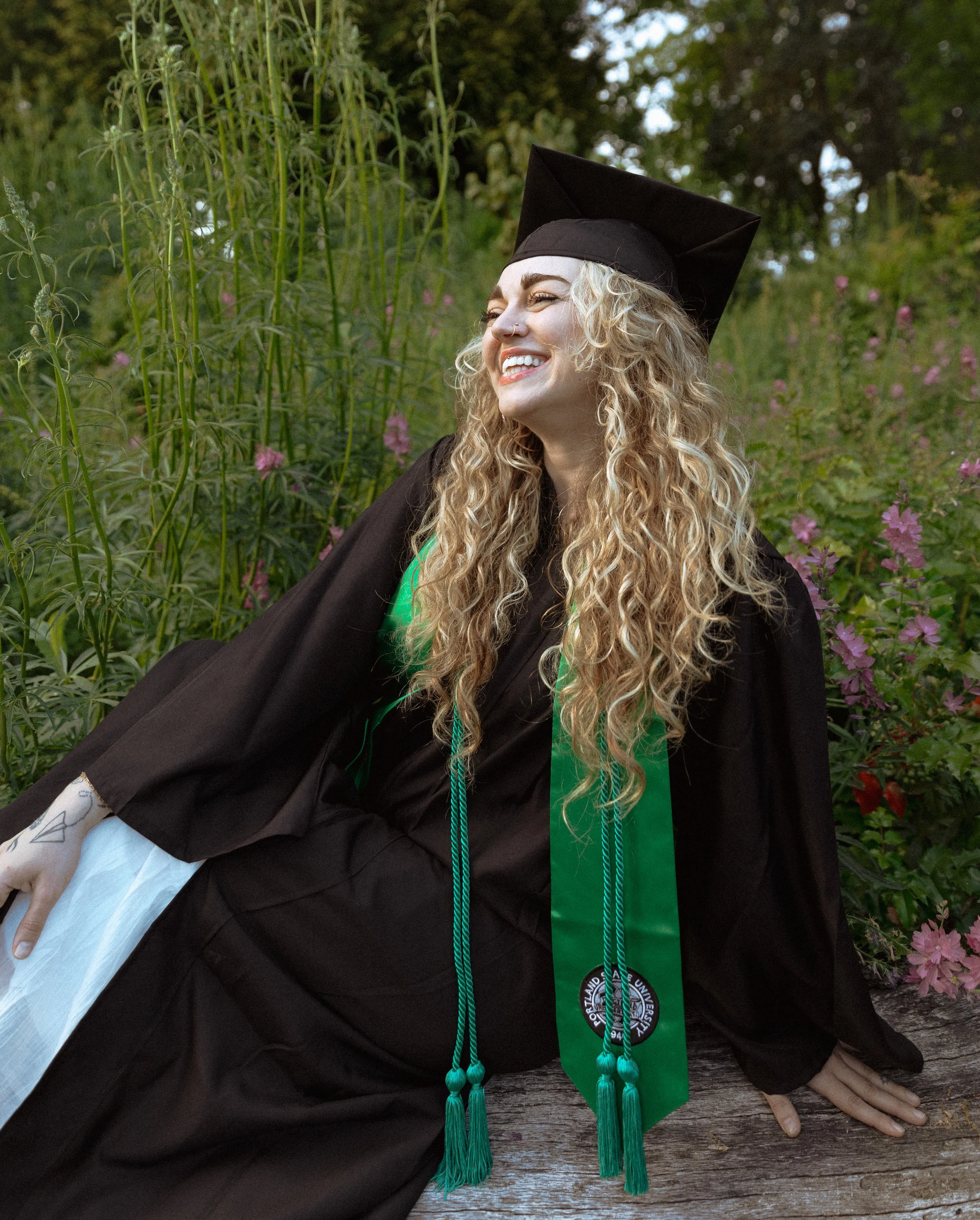 Portland University Grad with long curly blonde hair in graduation cap and gown, smiling, outdoors with green foliage and pink flowers in the background.