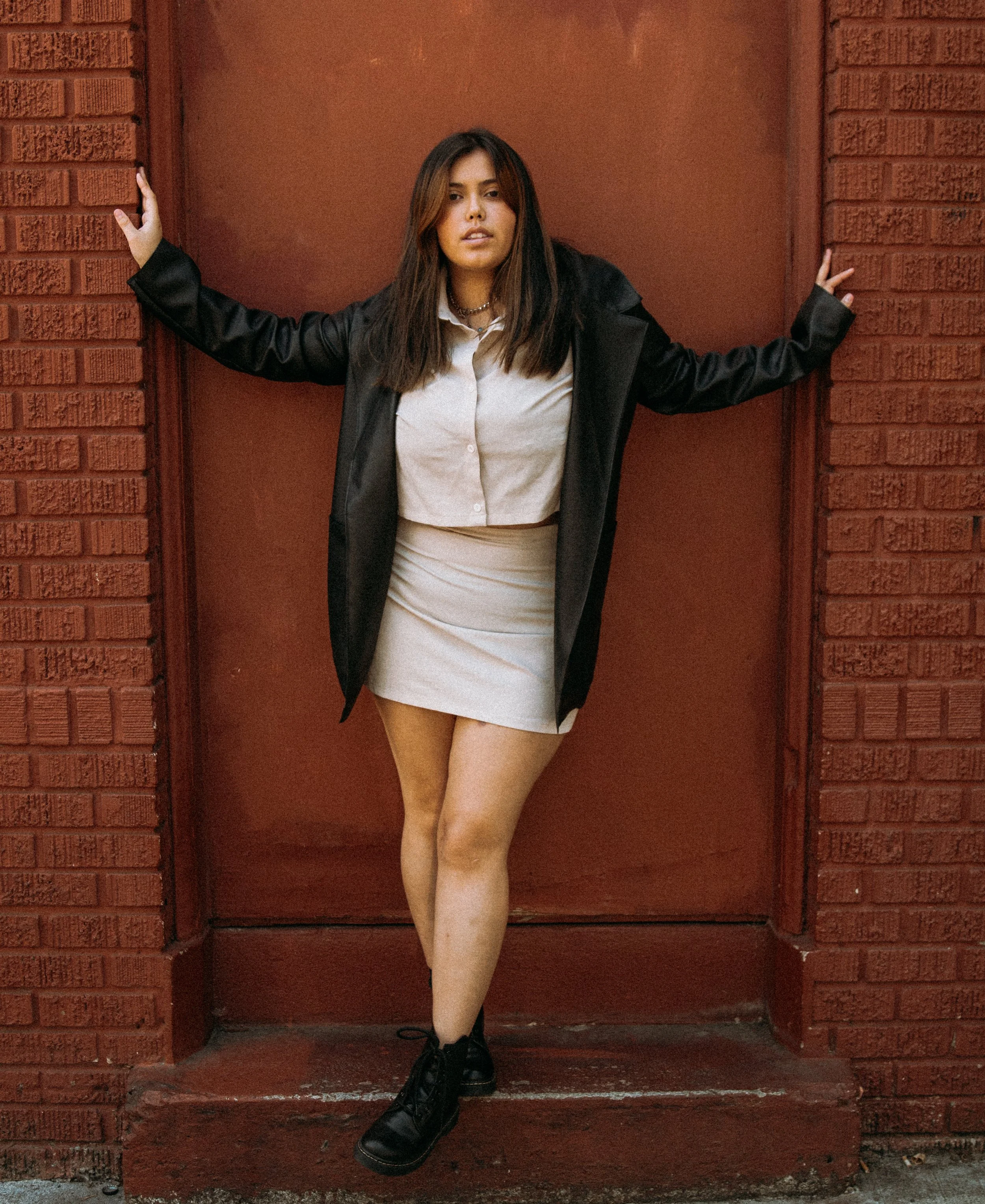 Woman with long brown hair wearing a white shirt, beige skirt, black jacket, and black boots, standing against a red brick wall with her arms outstretched, touching the wall on each side.