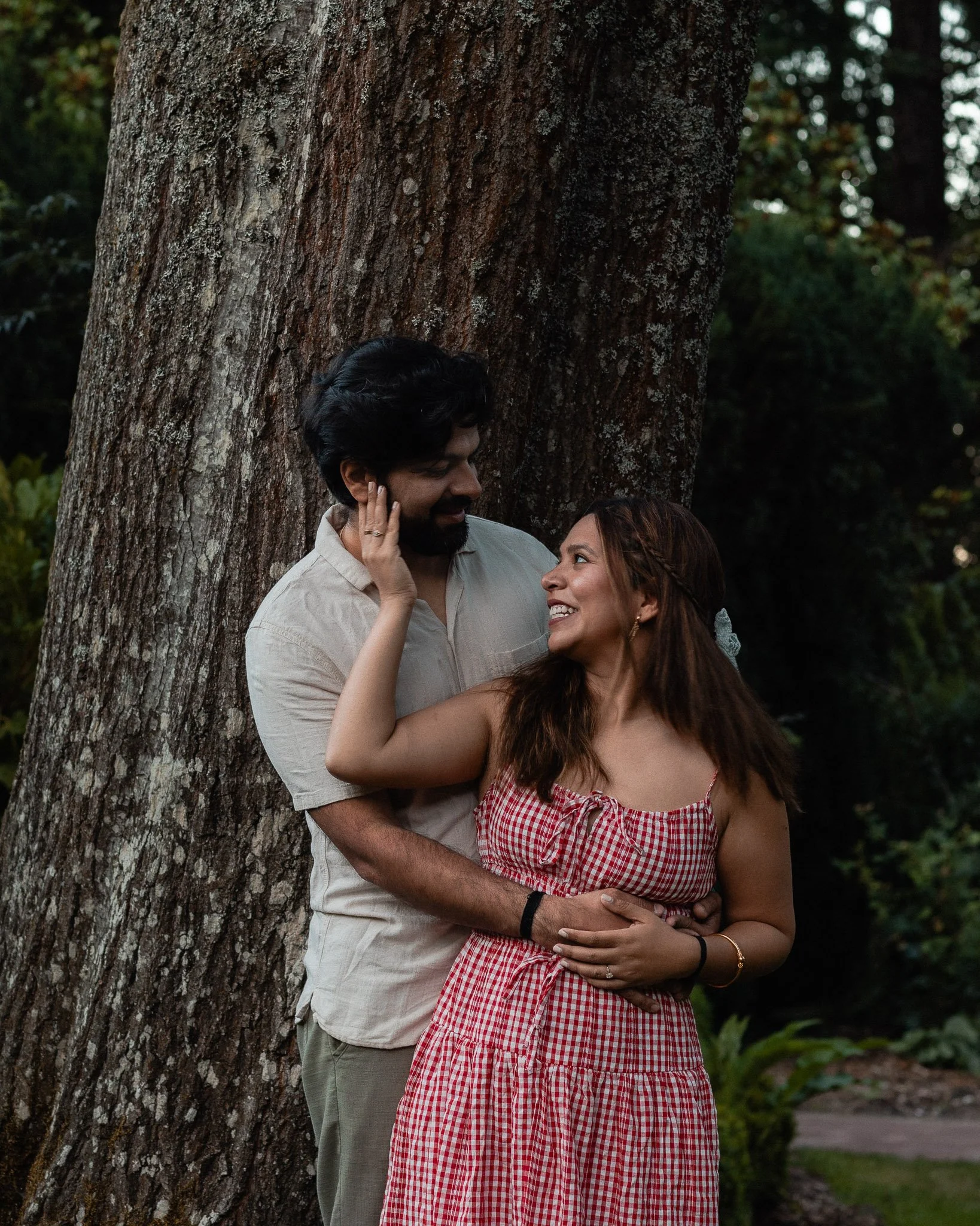 A man and woman standing close to each other outdoors, touching faces and gazing into each other's eyes near a large tree.
