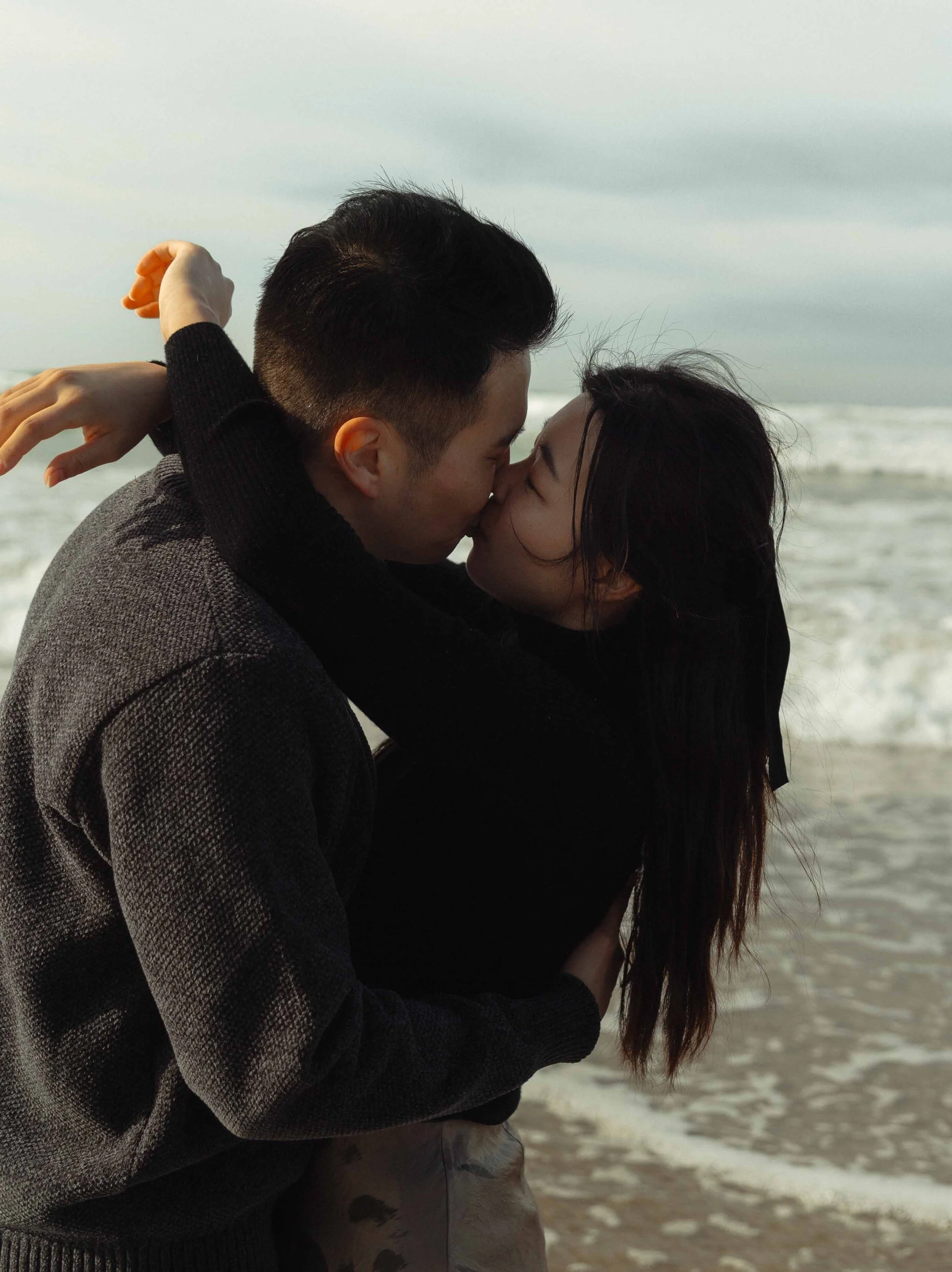 A couple kissing by the beach with waves in the background.