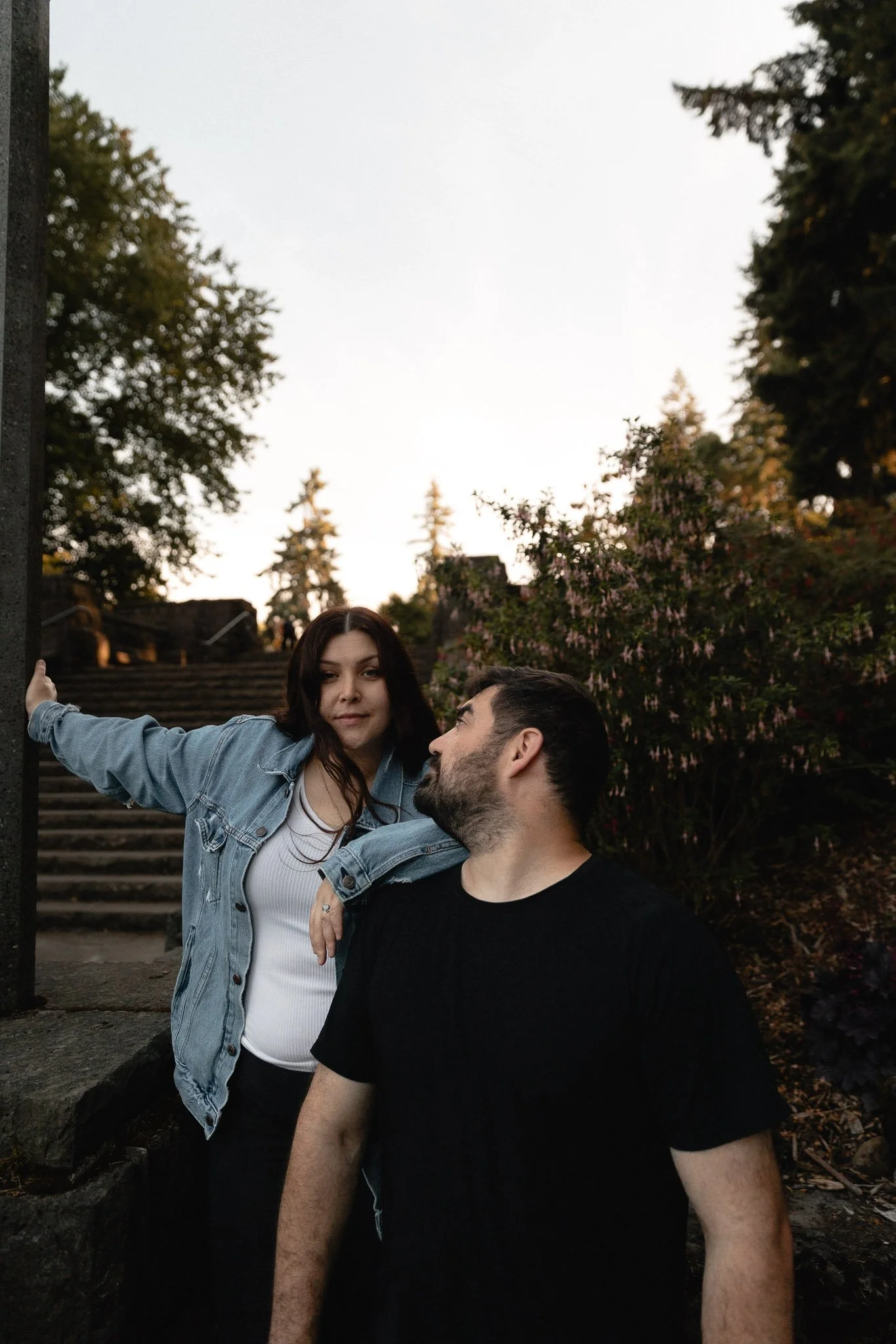 Two young adults, a woman and a man, standing outside near stairs with greenery and trees in the background during sunset. The woman is leaning on the man's shoulder, with her arm stretched out to the side, while the man is looking at her. The woman 