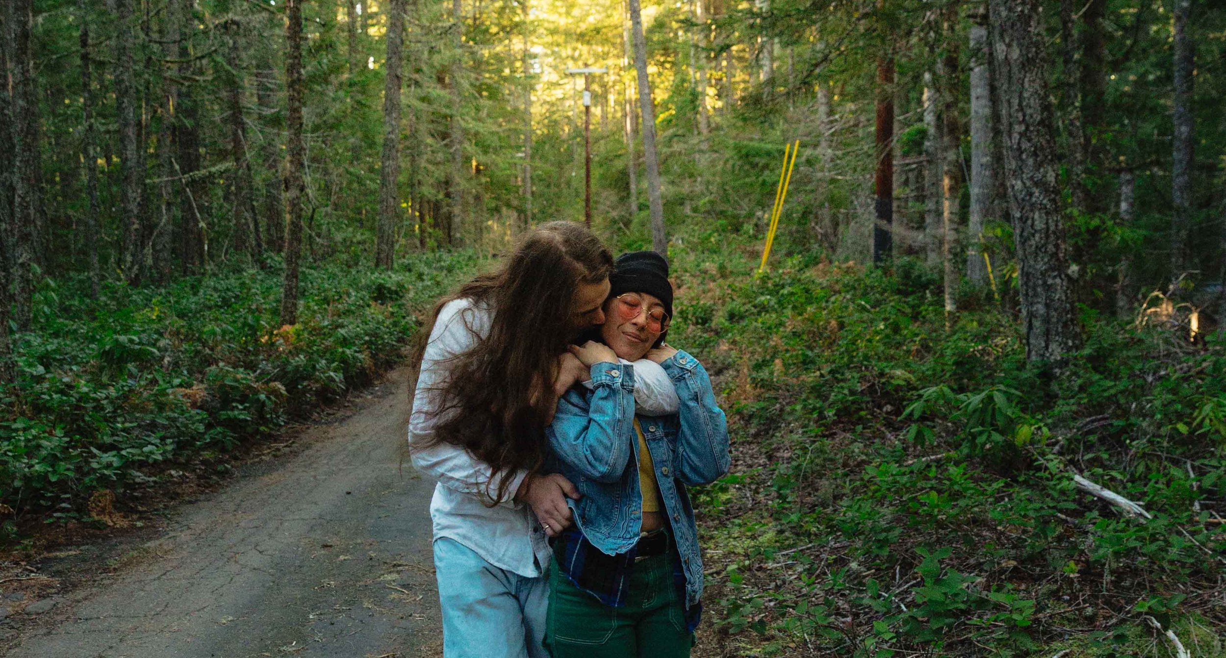 Two women walking in a forested trail, one comforting the other with a hug and a kiss on the cheek.