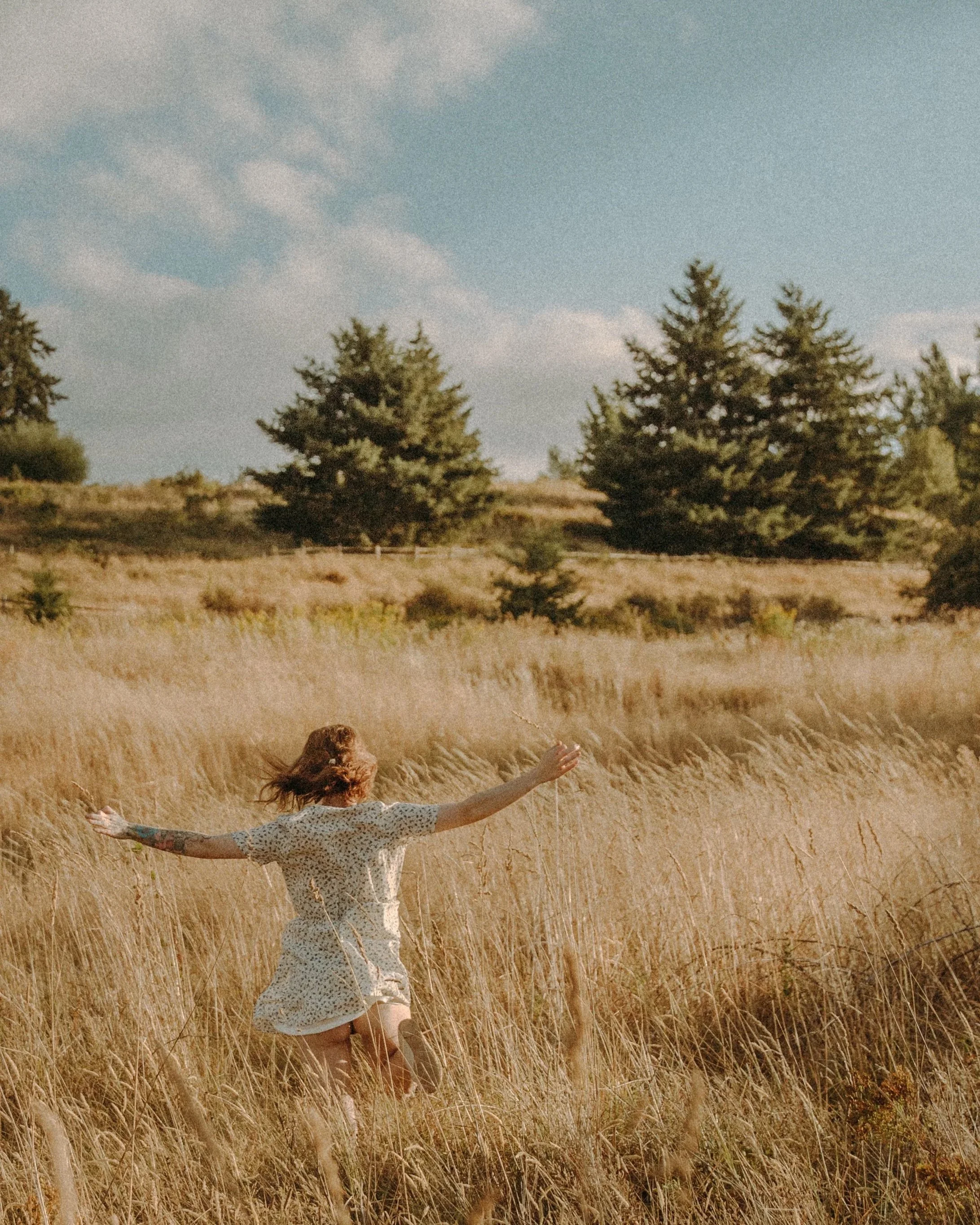A woman with tattoos on her arm, wearing a light-colored polka dot dress, is running through a golden wheat field with her arms outstretched. There are tall trees in the background and a blue sky with some clouds.