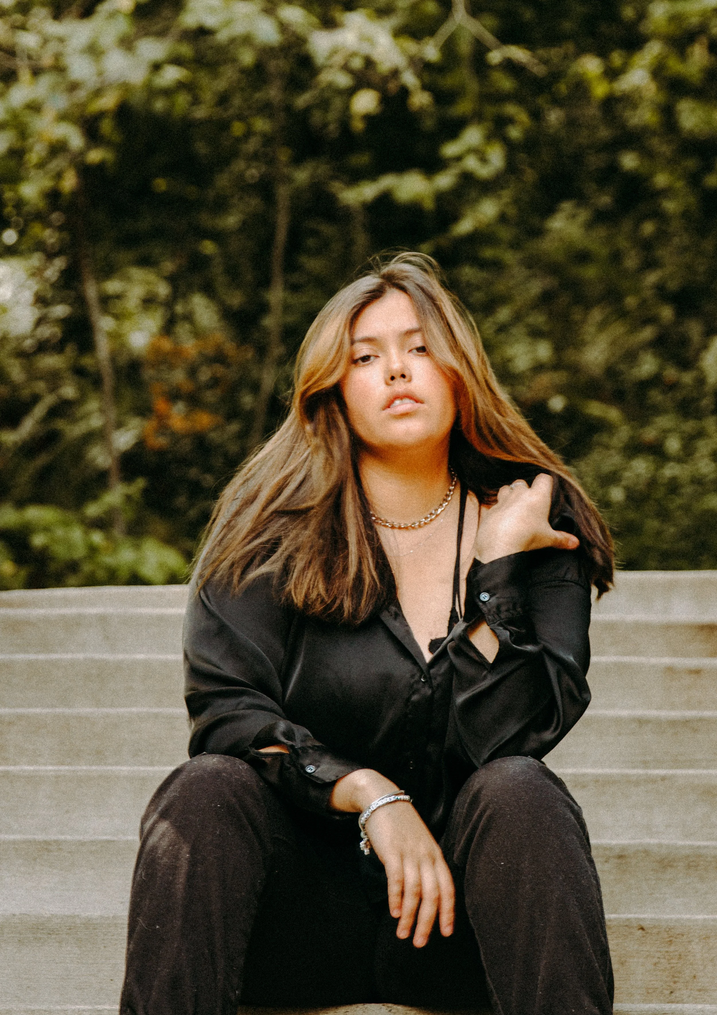 Young woman with brown hair, wearing black shirt and dark pants, sitting on outdoor stairs with a green, blurred background.