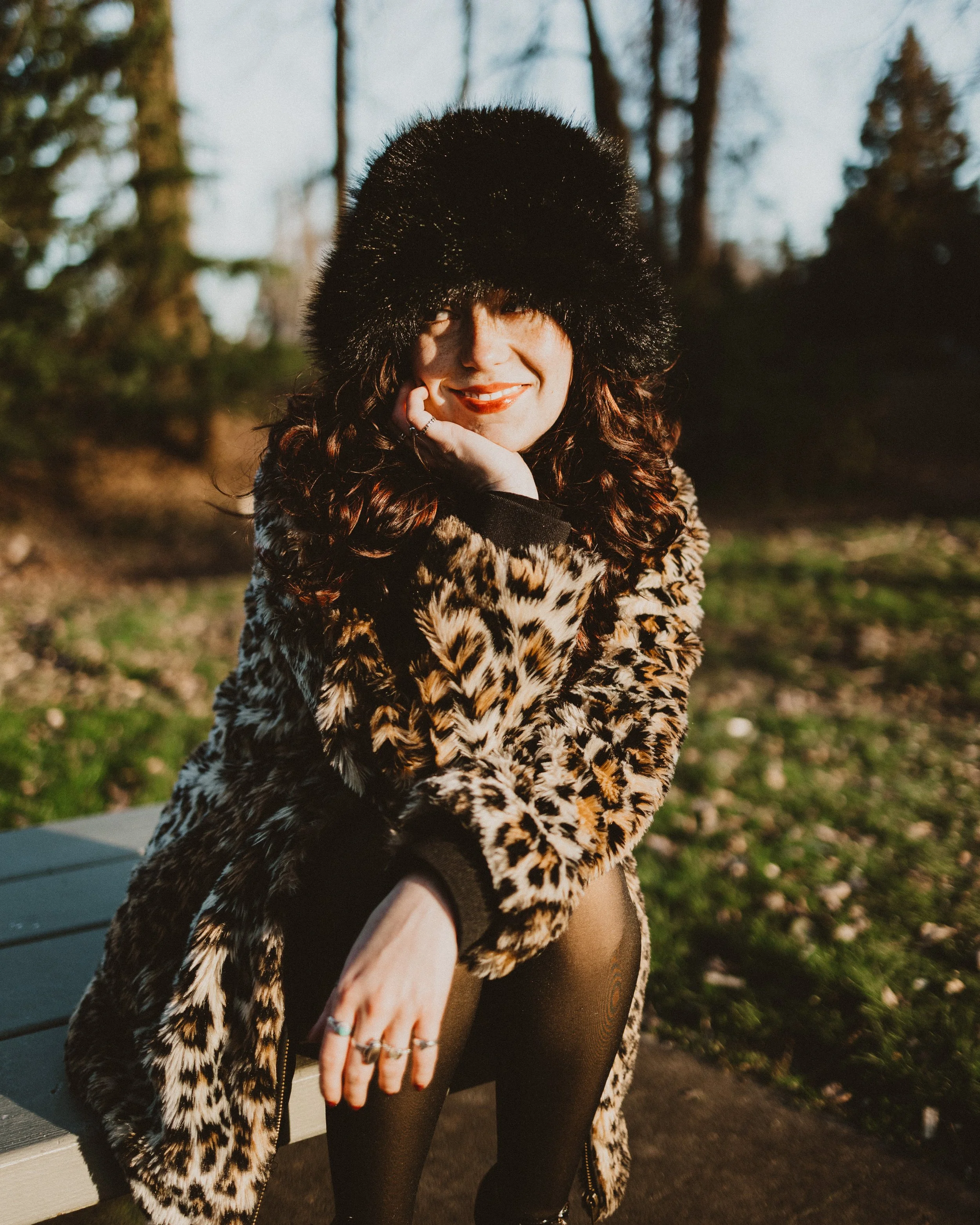 A woman wearing a black fur hat, a leopard print coat, and black tights standing outdoors during sunset, with her hand extended towards the camera.