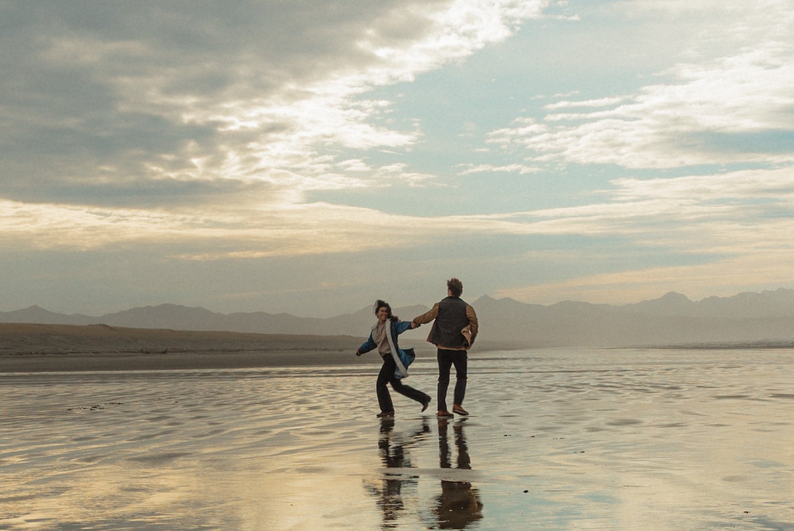 Two people holding hands and walking on a reflective wet beach with mountains and a partly cloudy sky in the background.