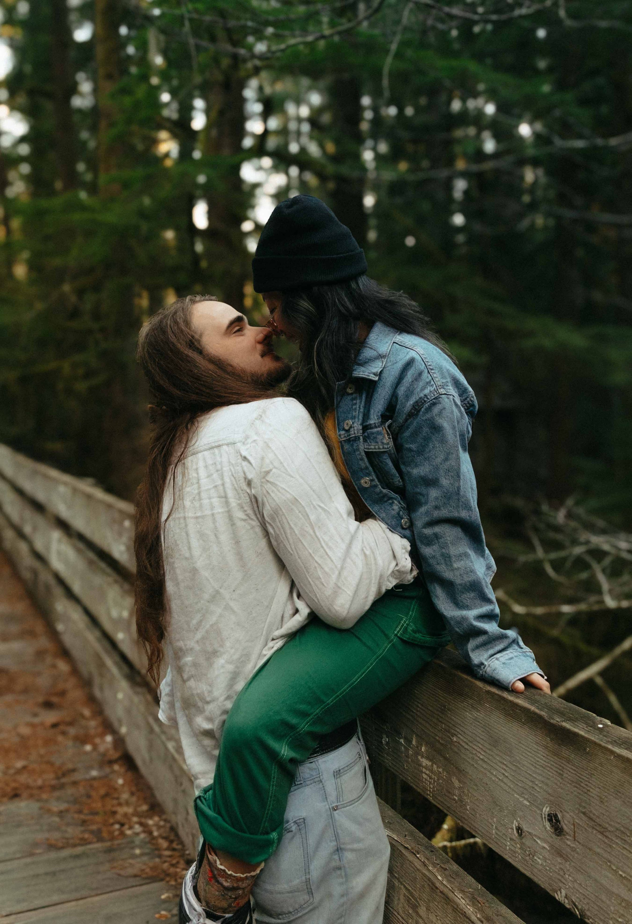 Two individuals are embracing outdoors on a wooden bridge surrounded by trees, with one person wearing a black beanie and jeans, and the other wearing a white shirt and green pants.
