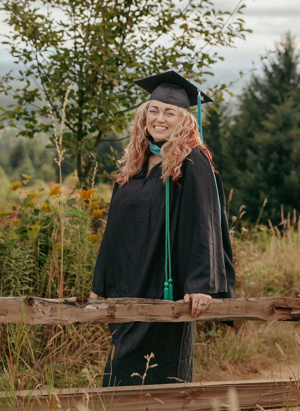 A woman in a black graduation cap and gown standing outdoors behind a wooden fence, smiling, with greenery and trees in the background.