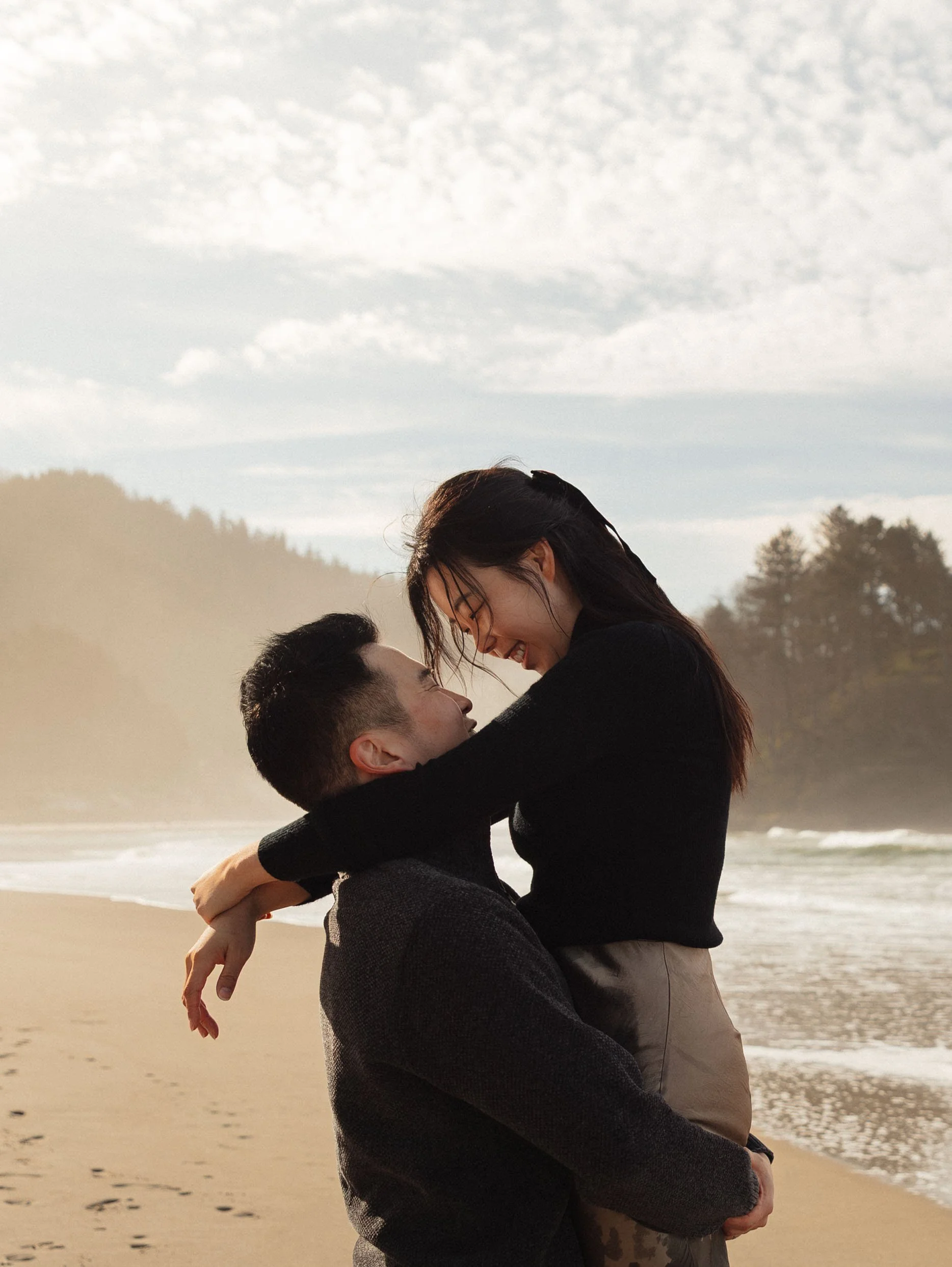 A happy couple is at the beach, with the woman being lifted by the man, smiling and looking at each other, near the water with a cloudy sky in the background.