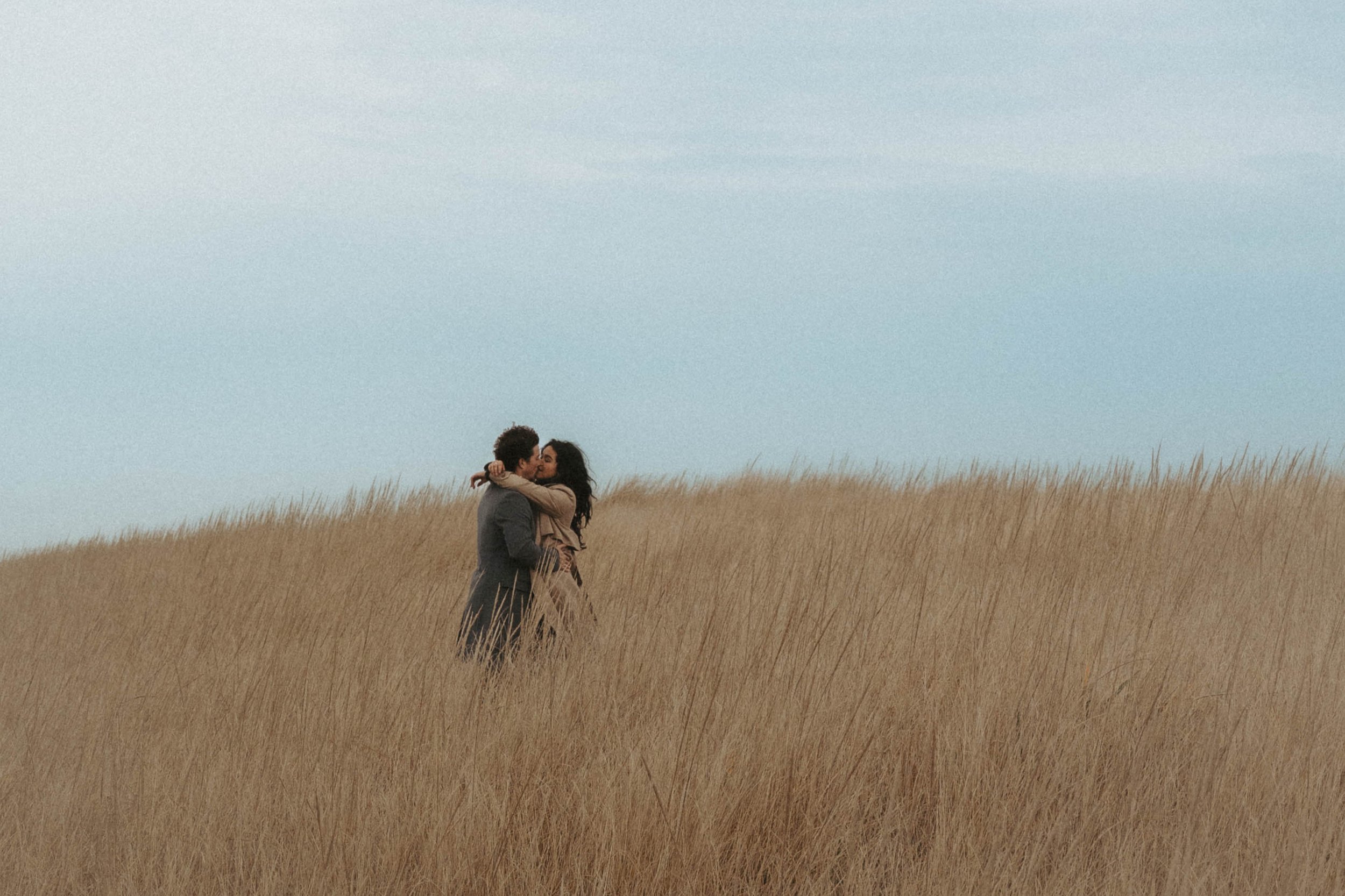 A couple embracing and kissing in a field of tall golden grass under a cloudy sky.