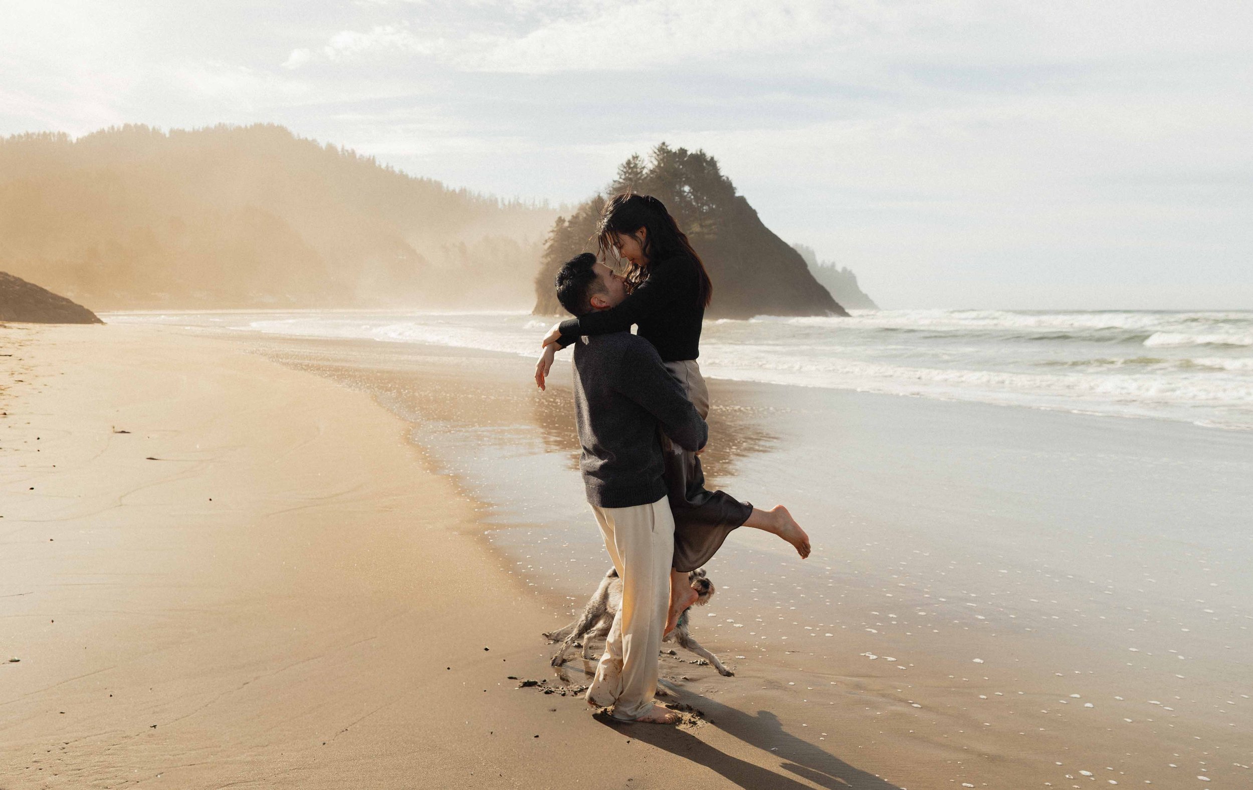 A couple on a beach, with the man lifting the woman in his arms and they are looking at each other, with rocky cliffs and the ocean in the background.