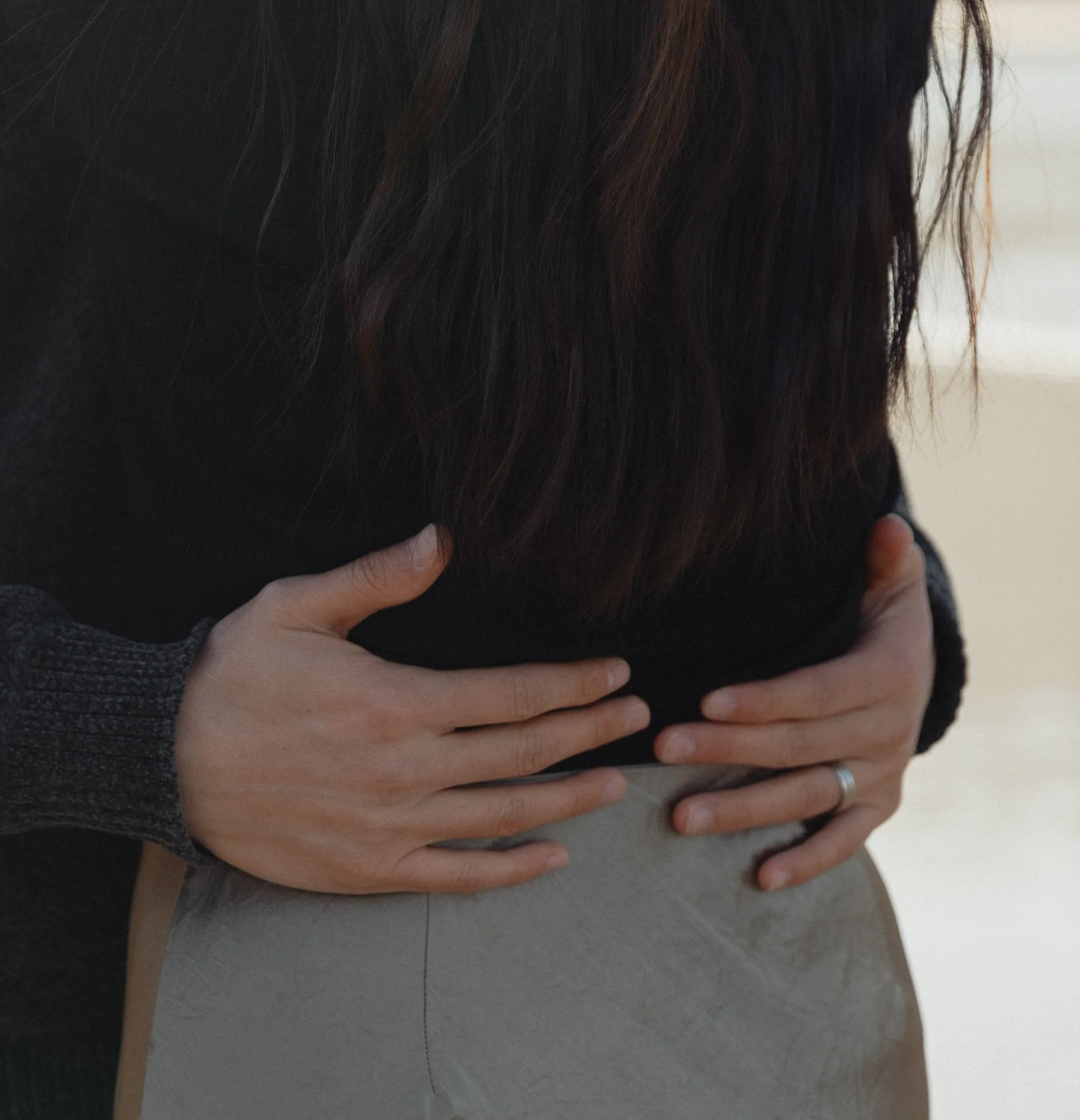 Close-up of two people holding hands, one with a wedding ring, against a neutral background.
