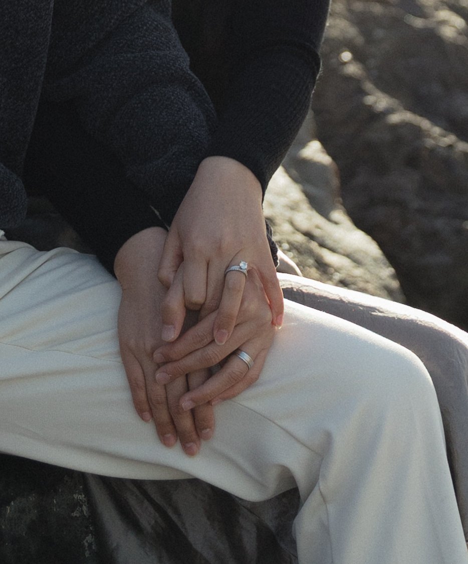 Close-up of two people holding hands, one wearing a ring, outdoors with rocky background.