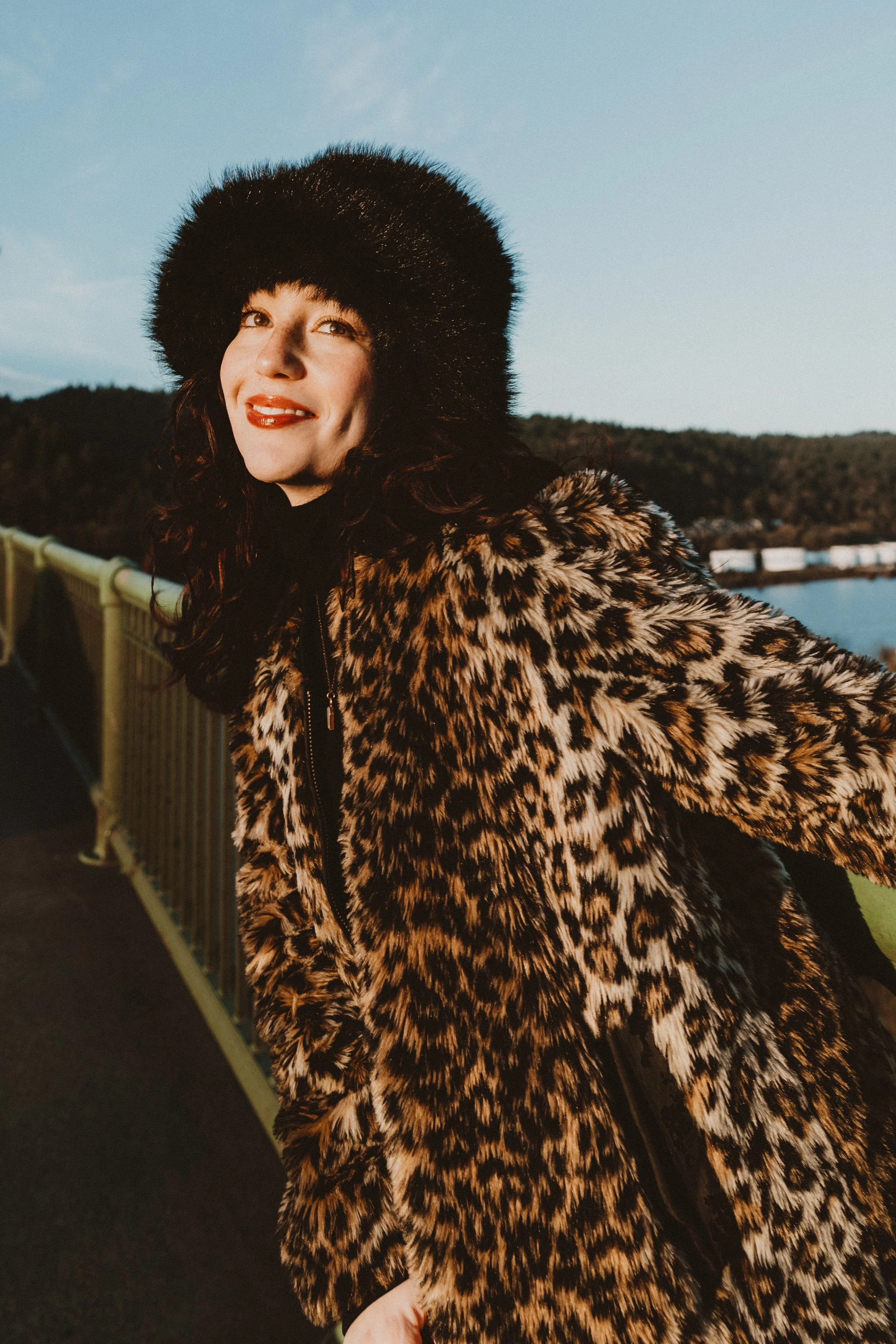 A woman smiling in a leopard print coat and a black furry hat standing outdoors near a body of water with hills in the background during early evening or late afternoon.
