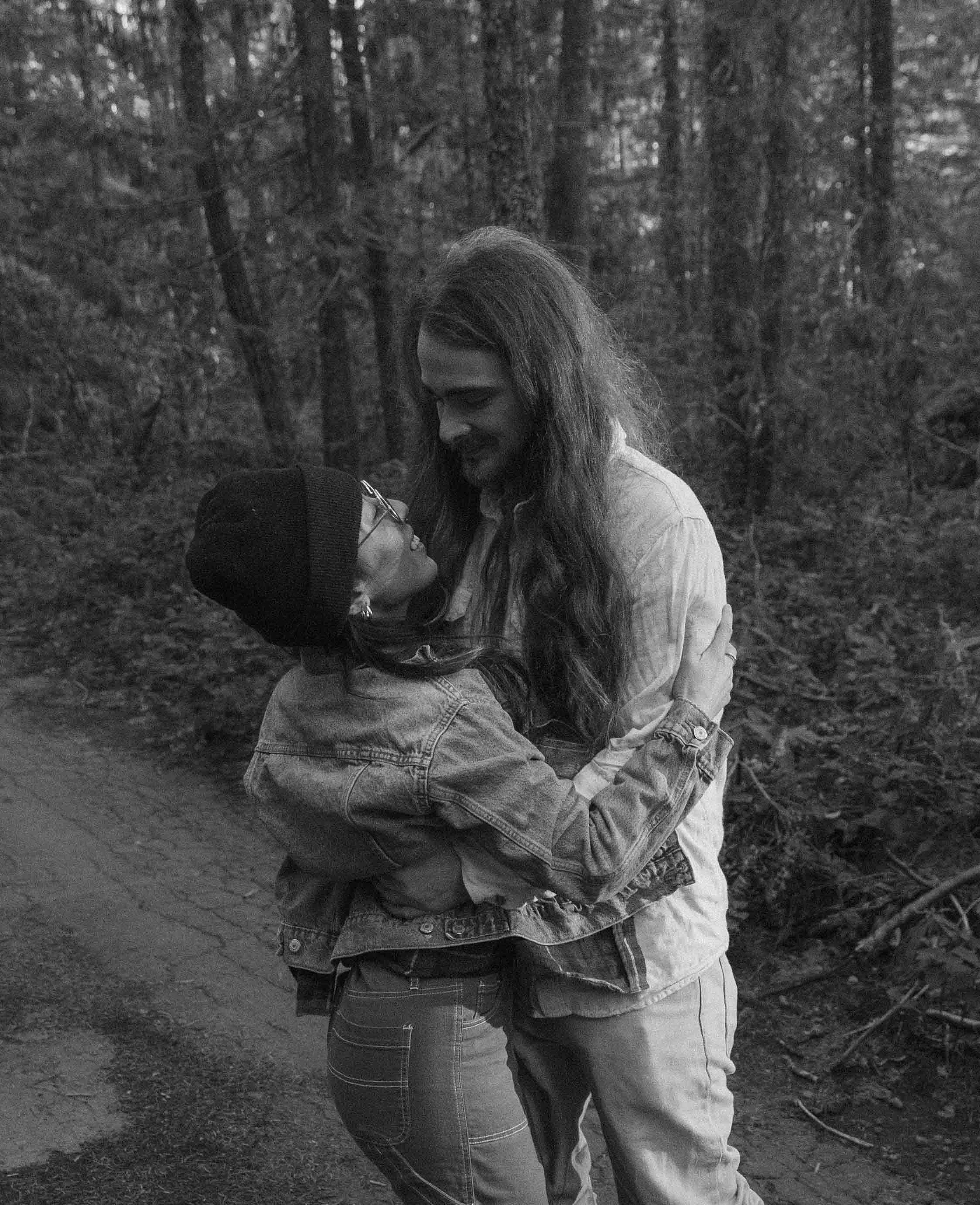 A black-and-white photo of a couple standing close together outdoors in a wooded area. The woman has long hair and is smiling, looking slightly downward. The man, wearing a beanie, glasses, and a jacket, is looking up towards the woman. They are embr