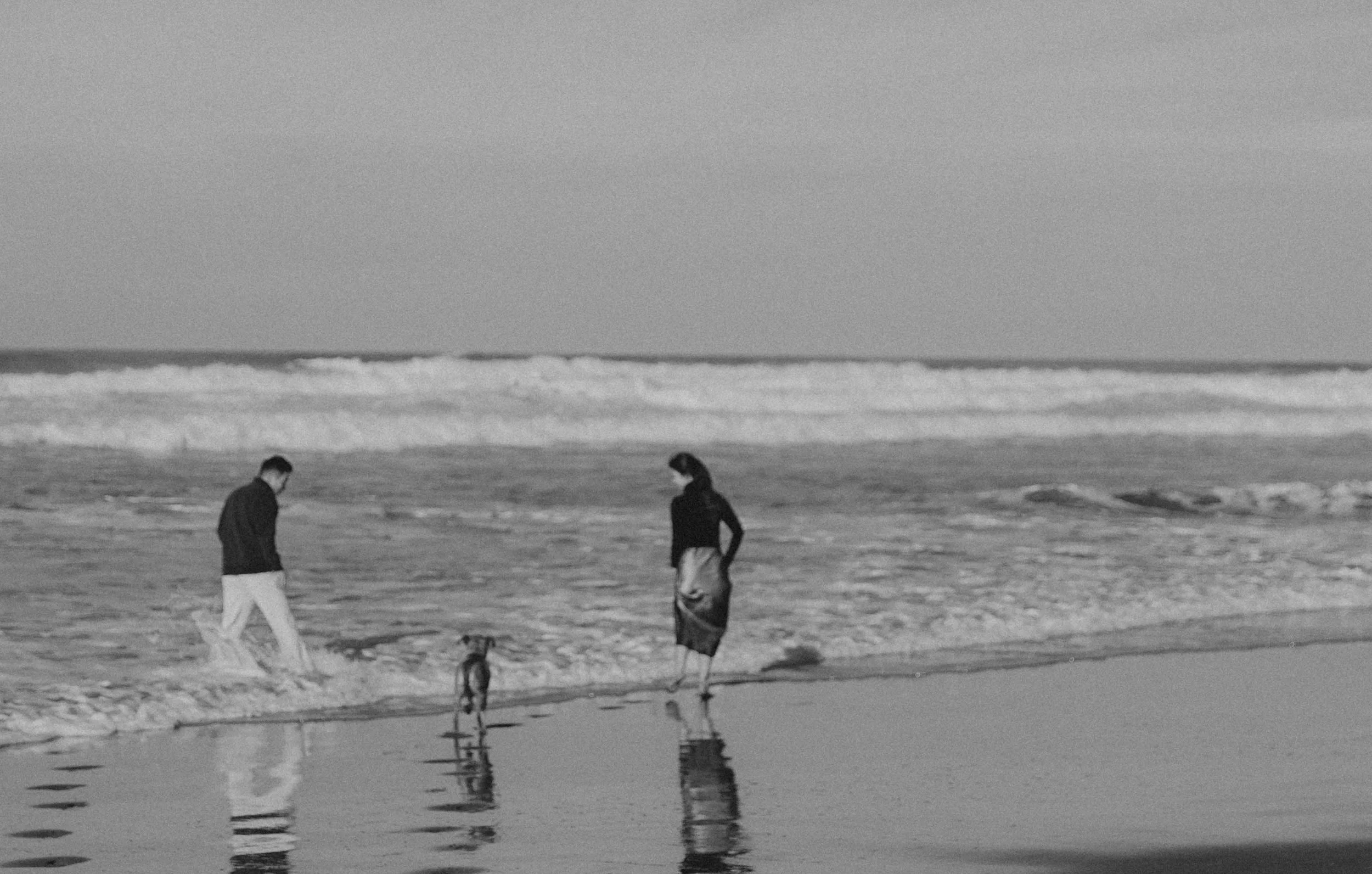 Three people on a beach, two walking and one standing, with waves in the background. Their reflections are visible in a small body of water in the foreground.