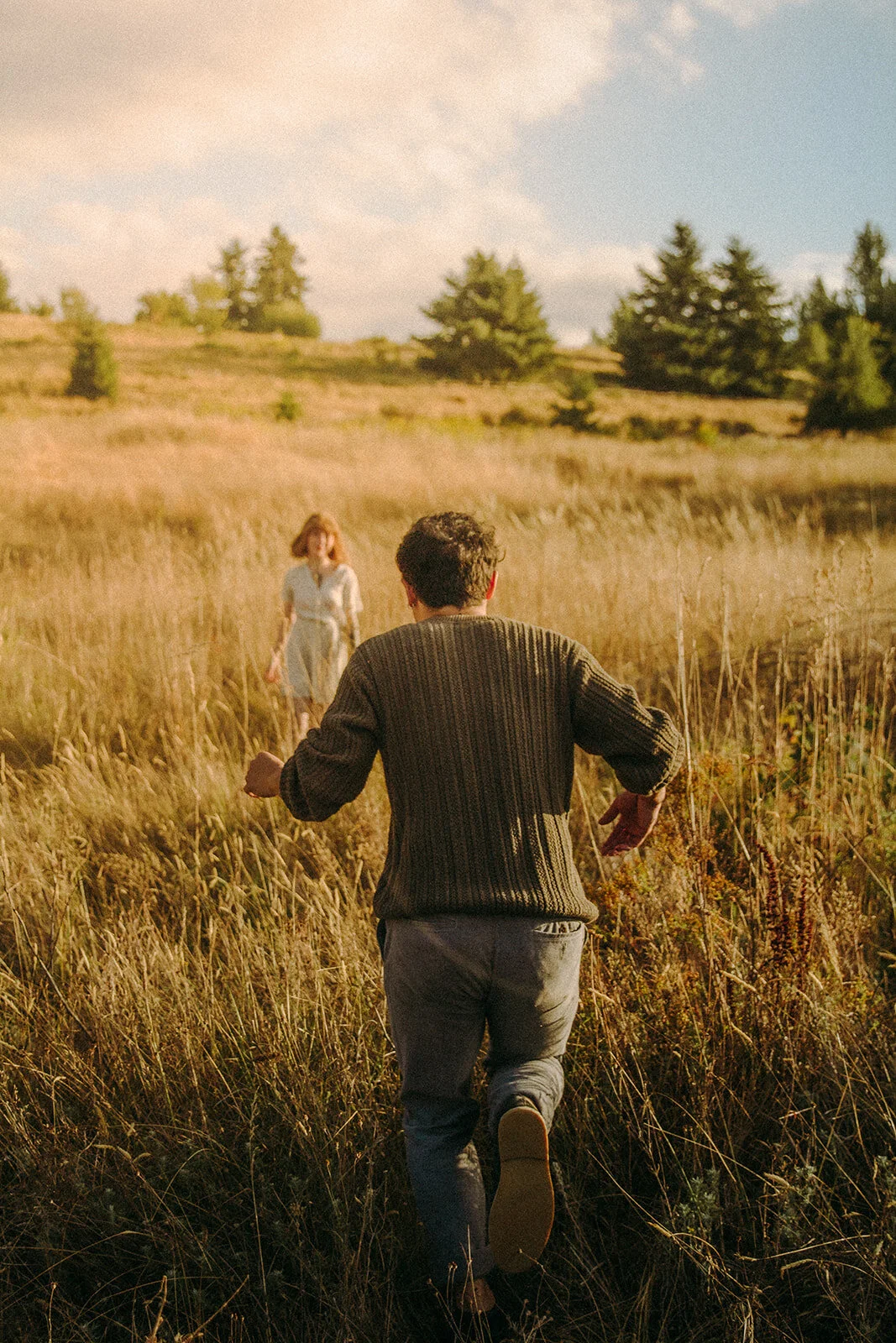 A man running through tall grass towards a woman in a field with trees and hills in the background.