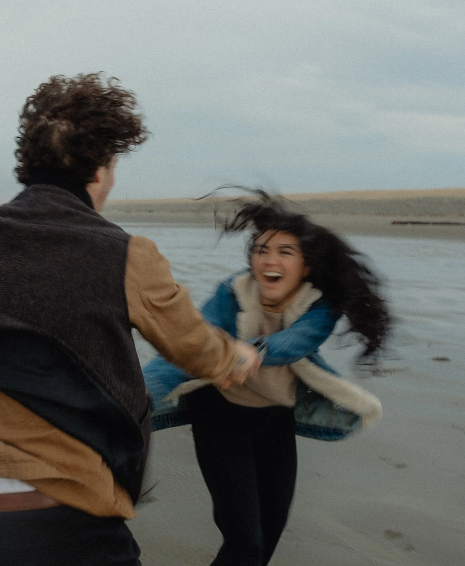 Two people, a man on the left and a woman on the right, are laughing and holding hands on a beach with water and sand in the background. The woman's hair is blowing in the wind.