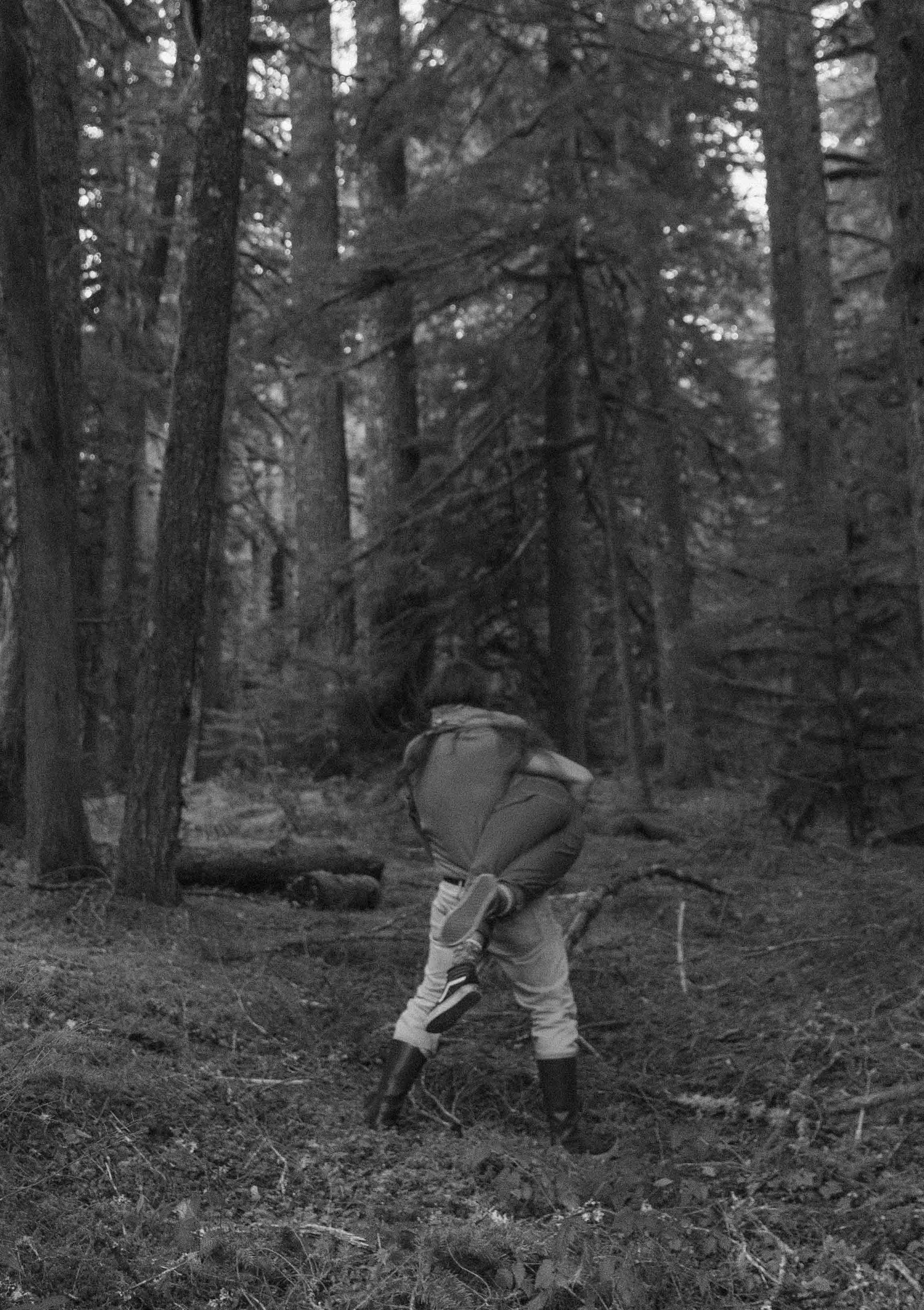 A child wearing a jacket and snow boots is kneeling on one knee in a dense forest of tall trees with sparse branches, looking upward.