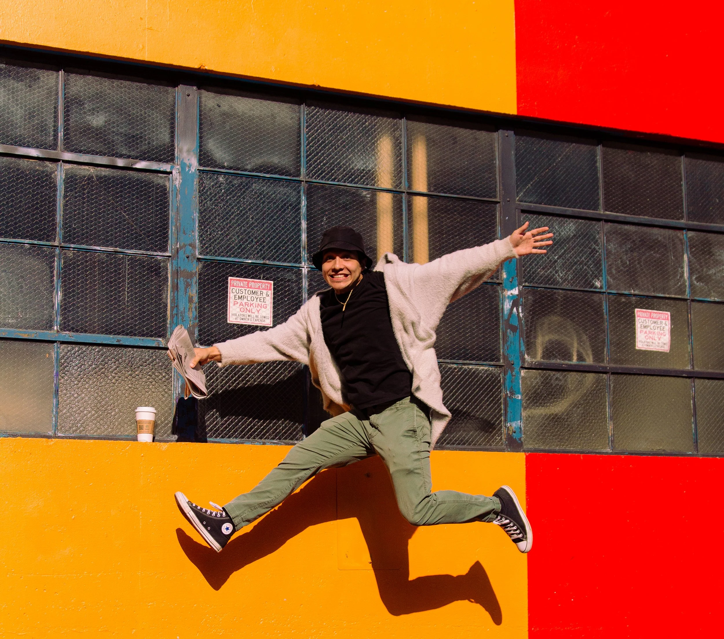 A person with a big smile, wearing a bucket hat, black shirt, and beige jacket, jumping with arms and legs extended against a colorful wall with orange, yellow, and red sections.