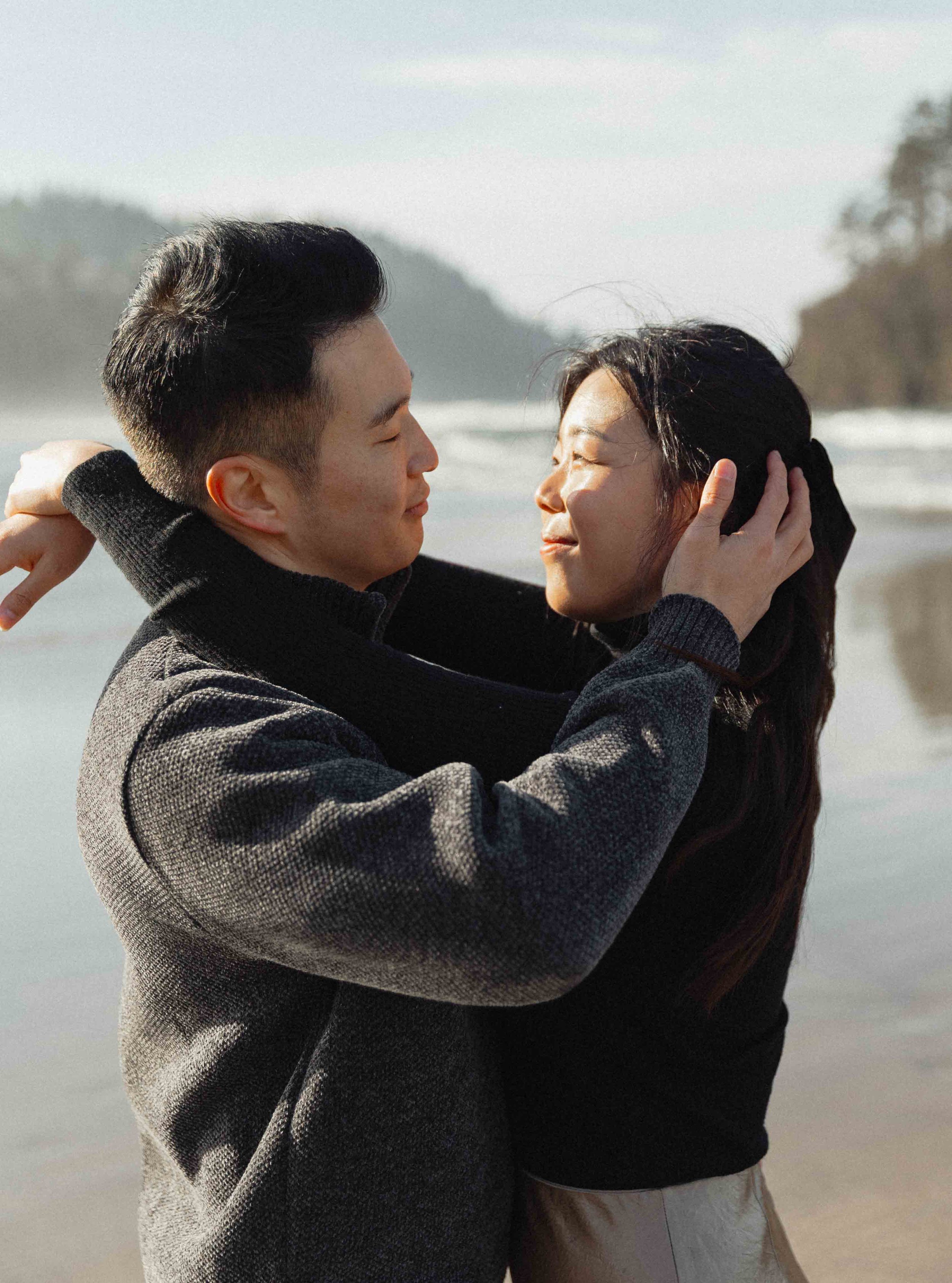 A close-up of a young couple embracing by the beach, with the man holding the woman's face, and the ocean and hazy hills in the background.