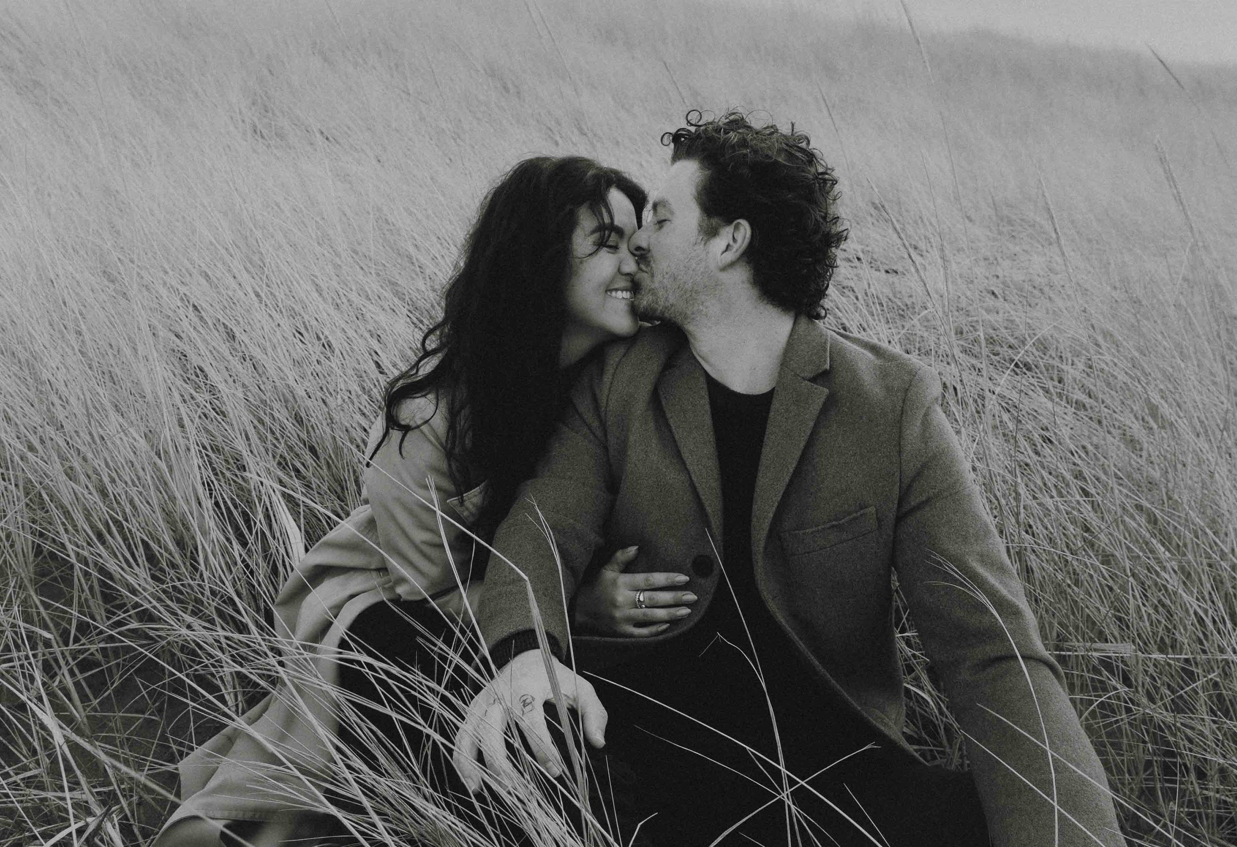 A couple sitting in tall grass, touching foreheads, smiling, in black-and-white photograph.